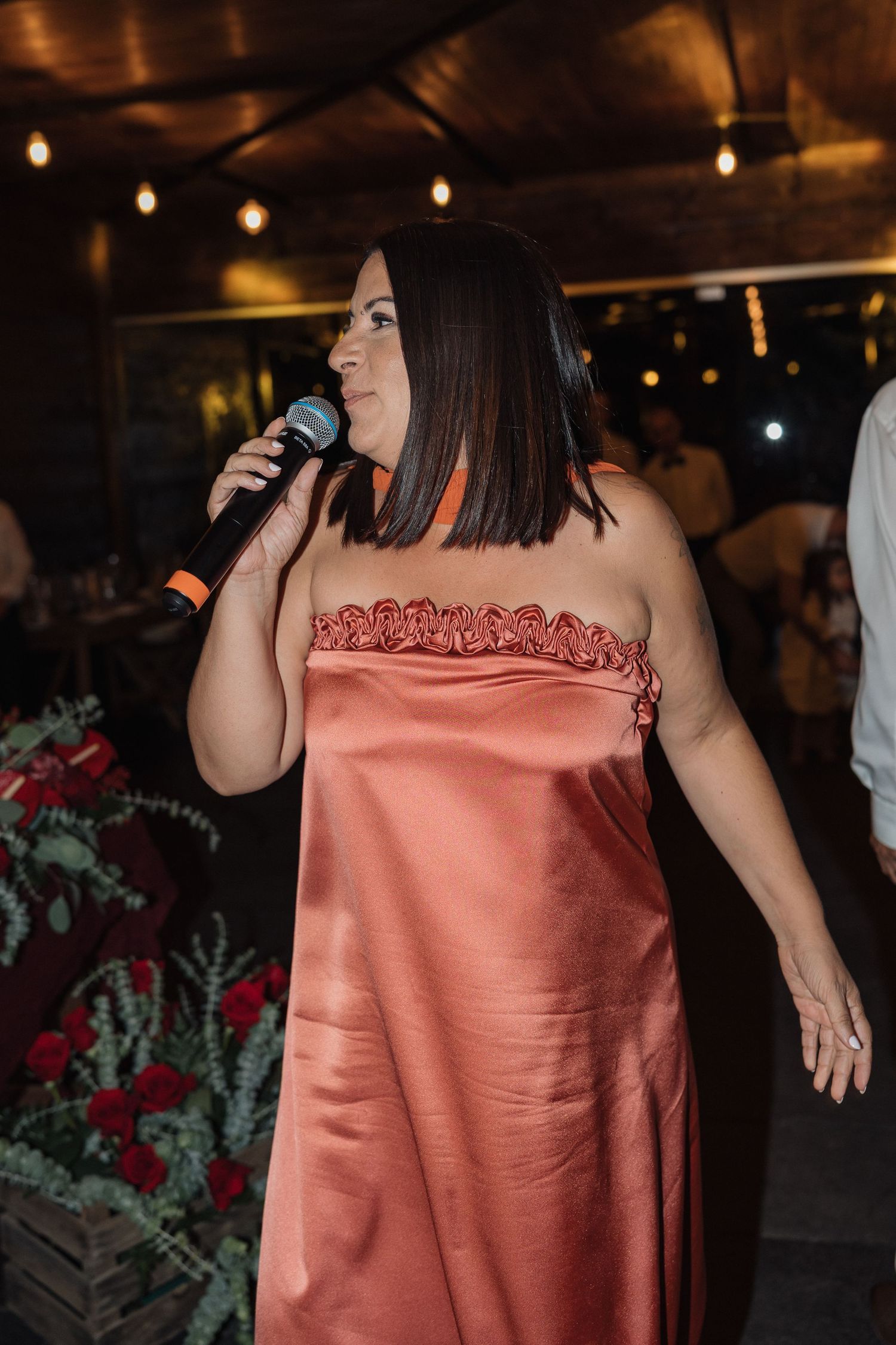 A performer in a coral satin dress sings at an evening event under string lights.