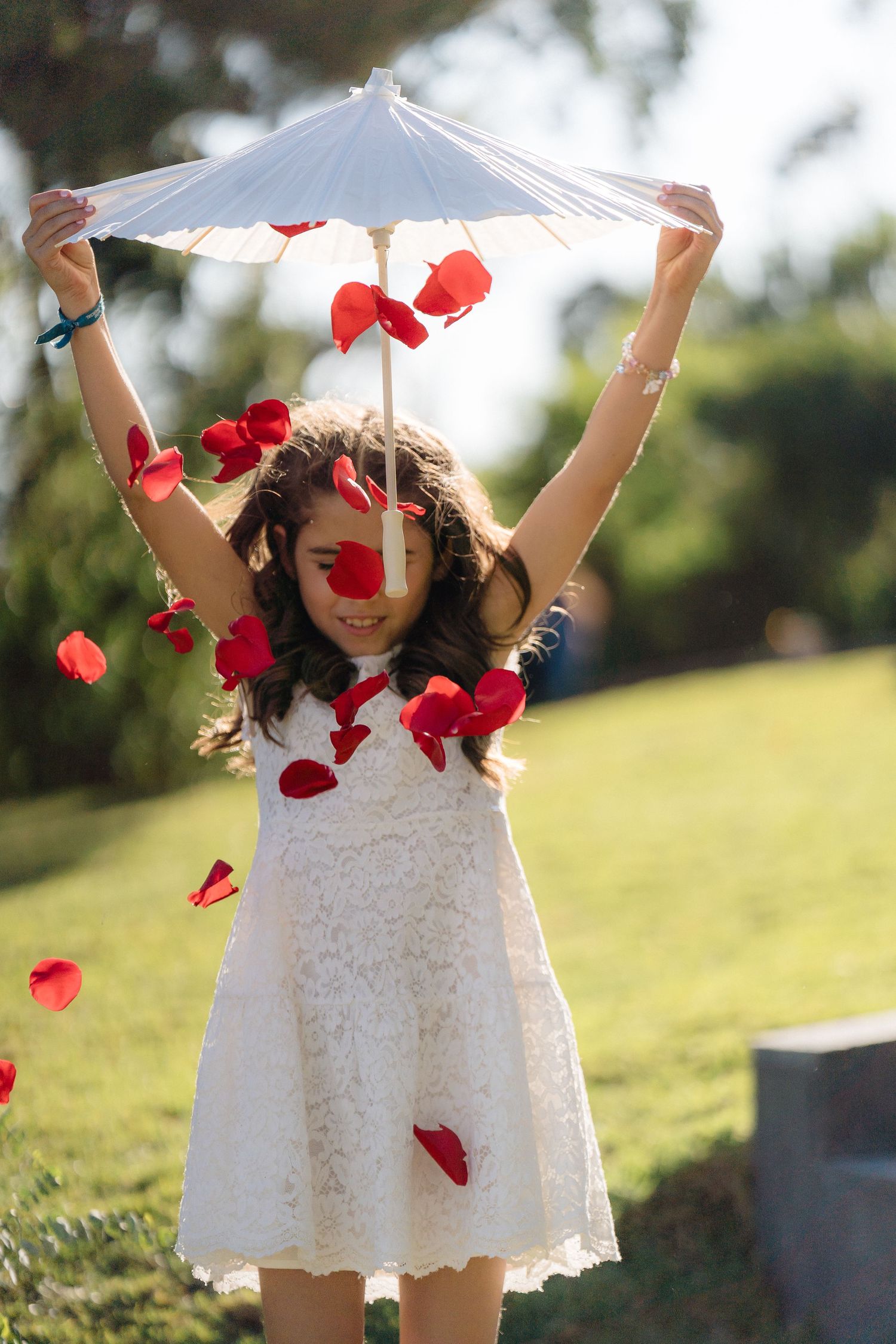 Young flower girl in white dress playing with parasol and red rose petals.