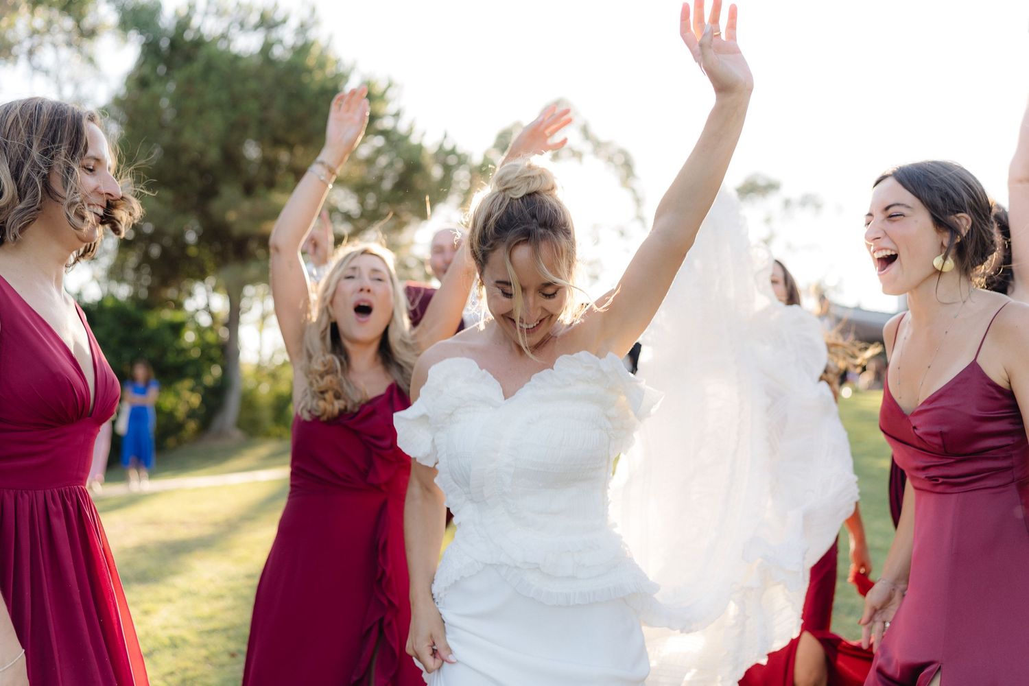 Celebratory moment at outdoor wedding with attendees in burgundy dresses dancing and laughing.