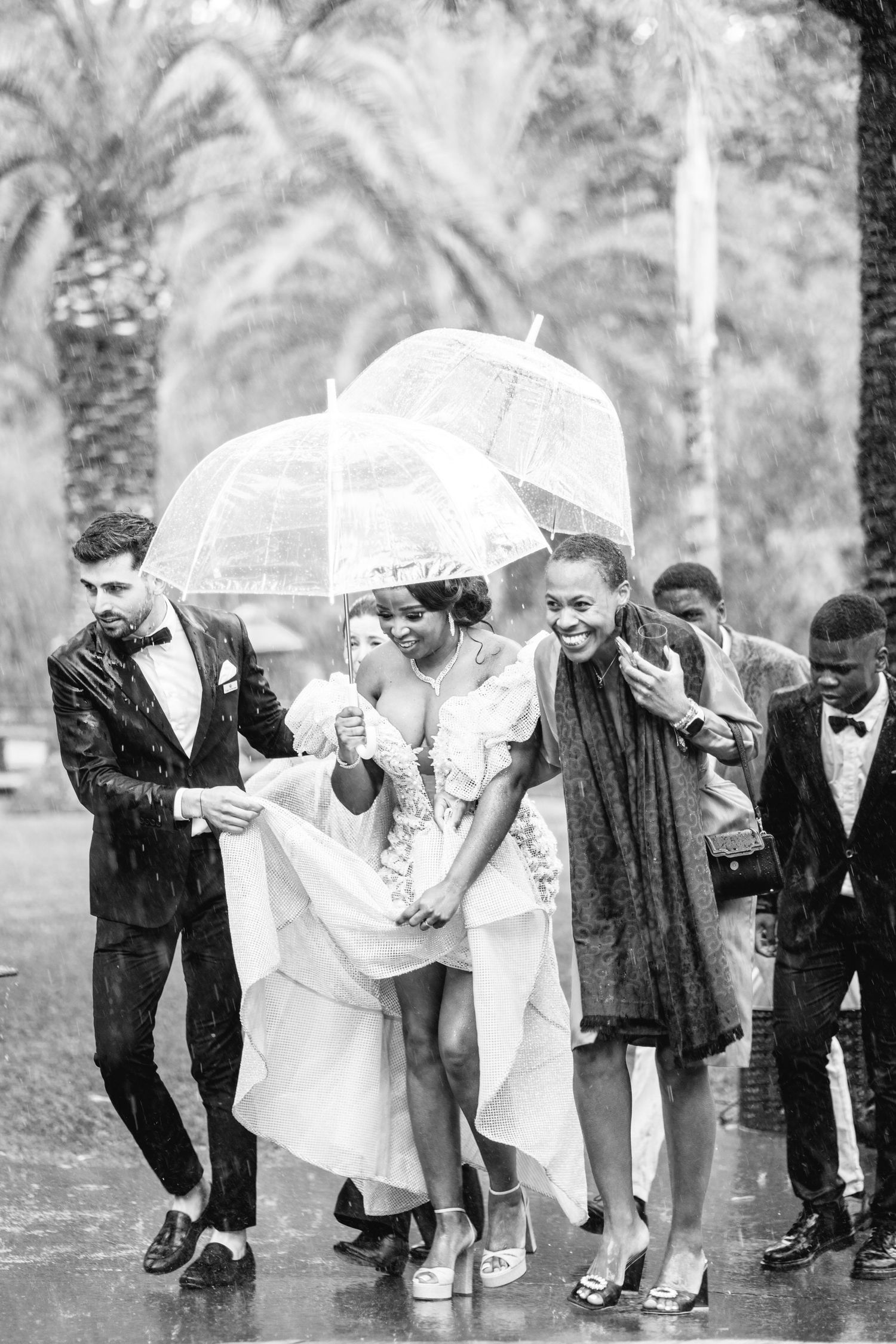 Wedding party huddles under white umbrellas during a rainy celebration outdoors.