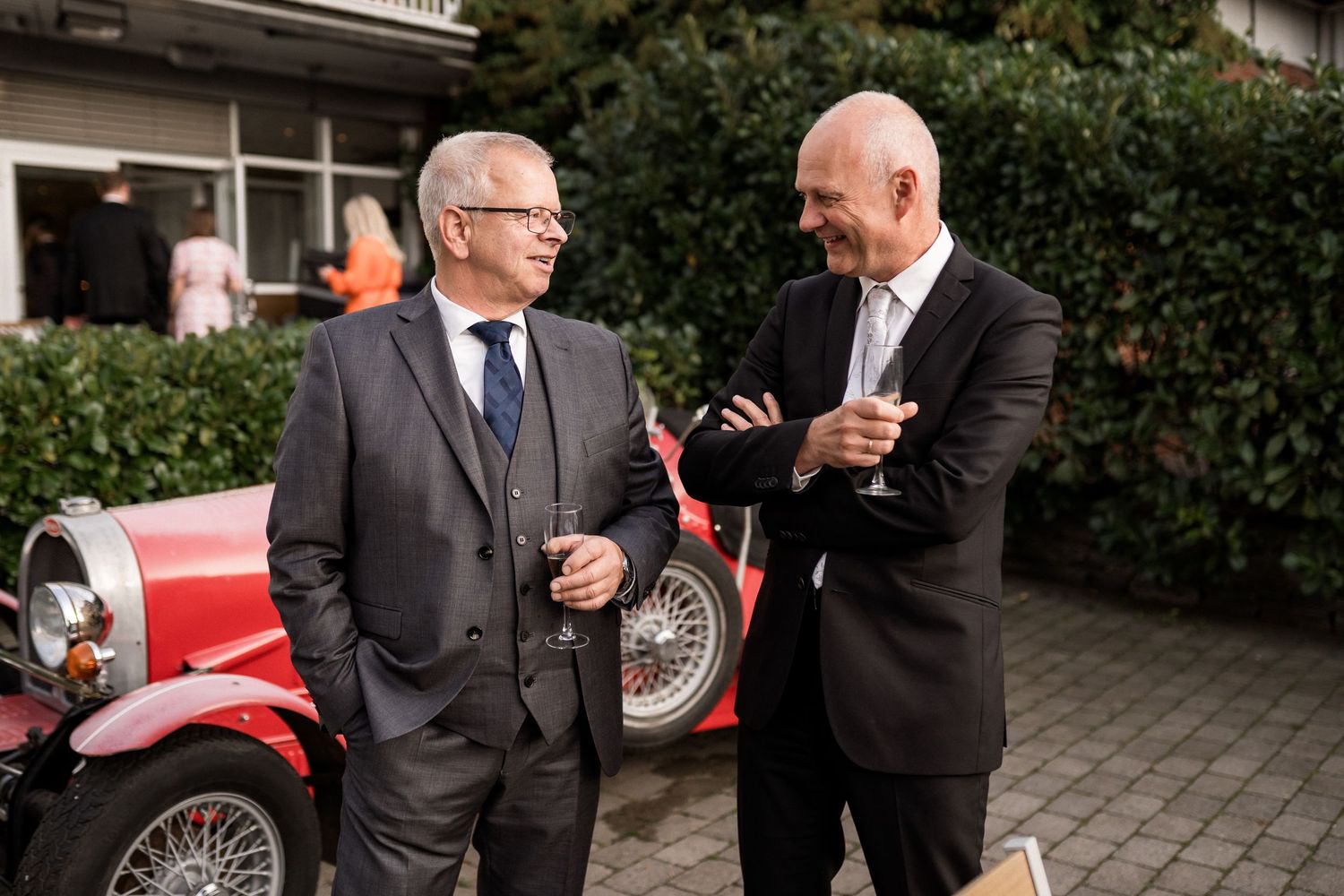 Wedding guests in formal attire gather around classic cars during an outdoor celebration.