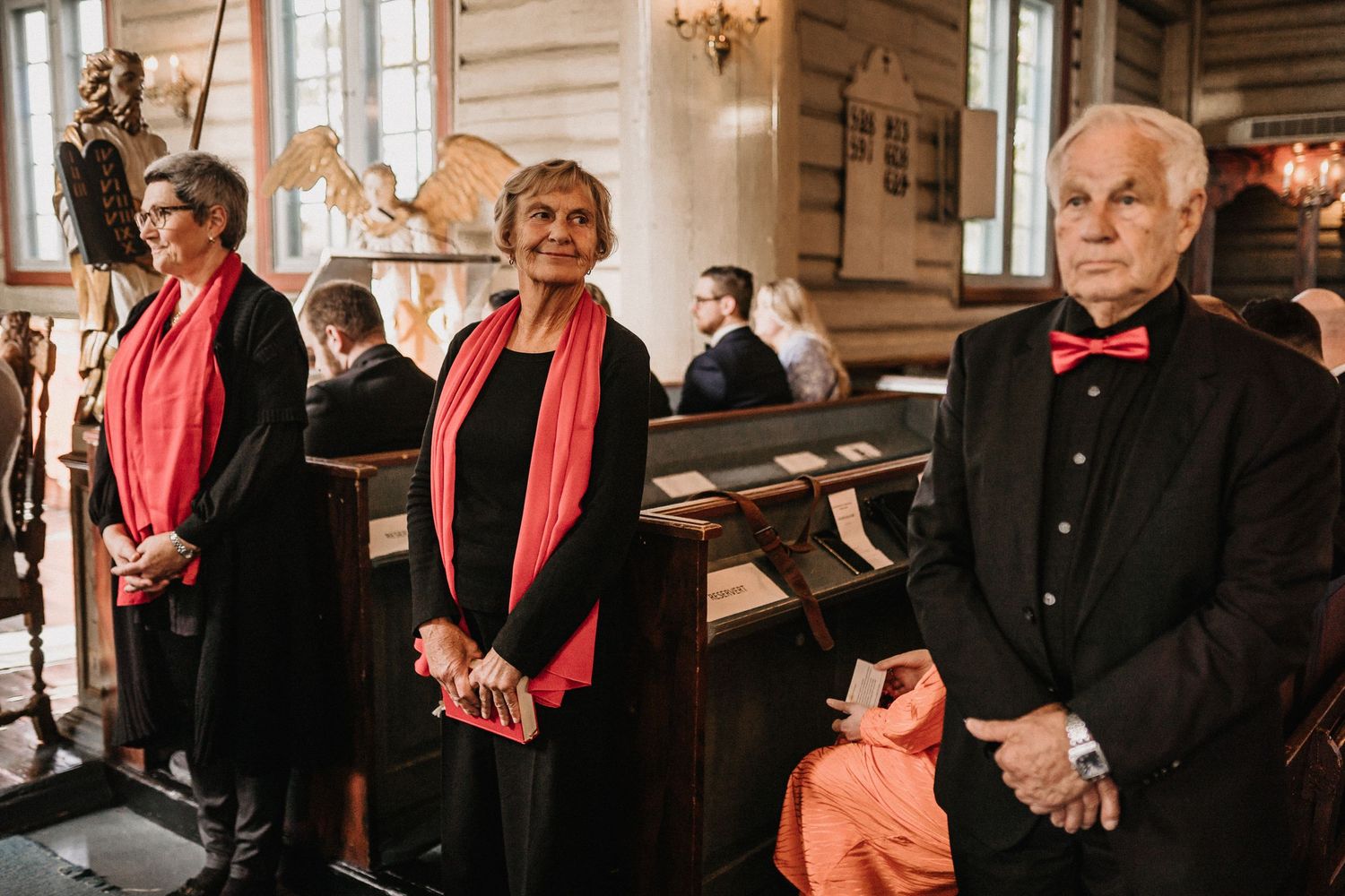 Choir members wearing red scarves stand together in a traditional church setting during a performance.