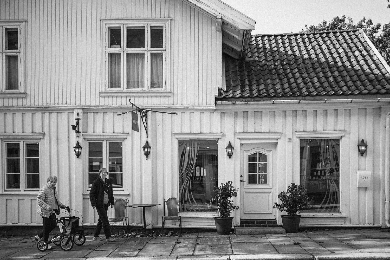 Panoramic black and white exterior view of a traditional white wooden building with multiple windows and wall lamps.