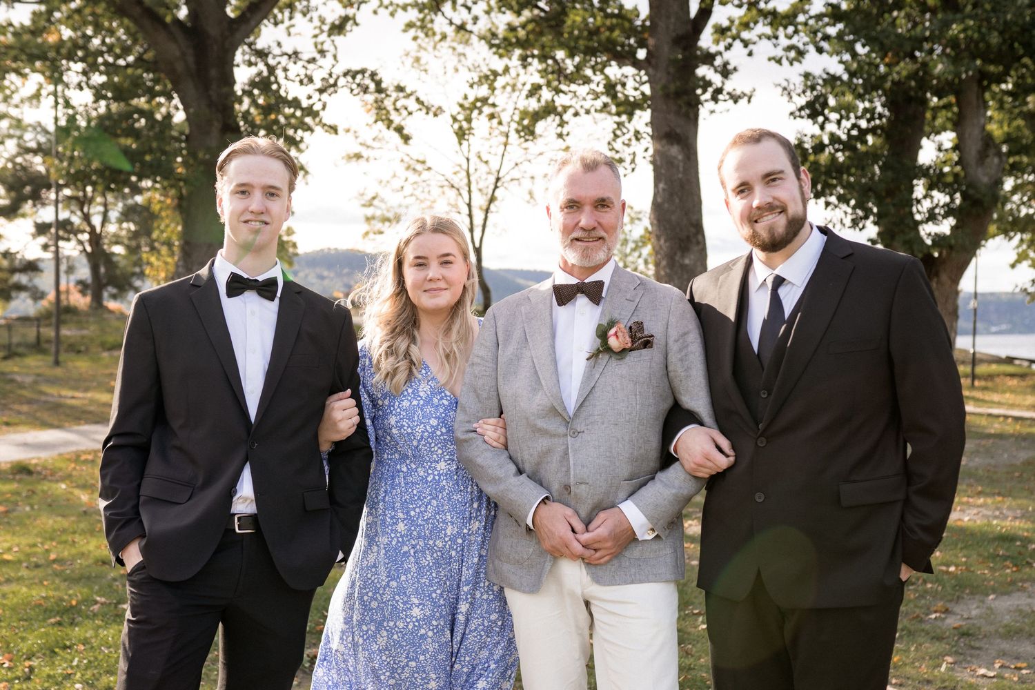 Wedding party poses outdoors under trees wearing formal attire and blue dresses.