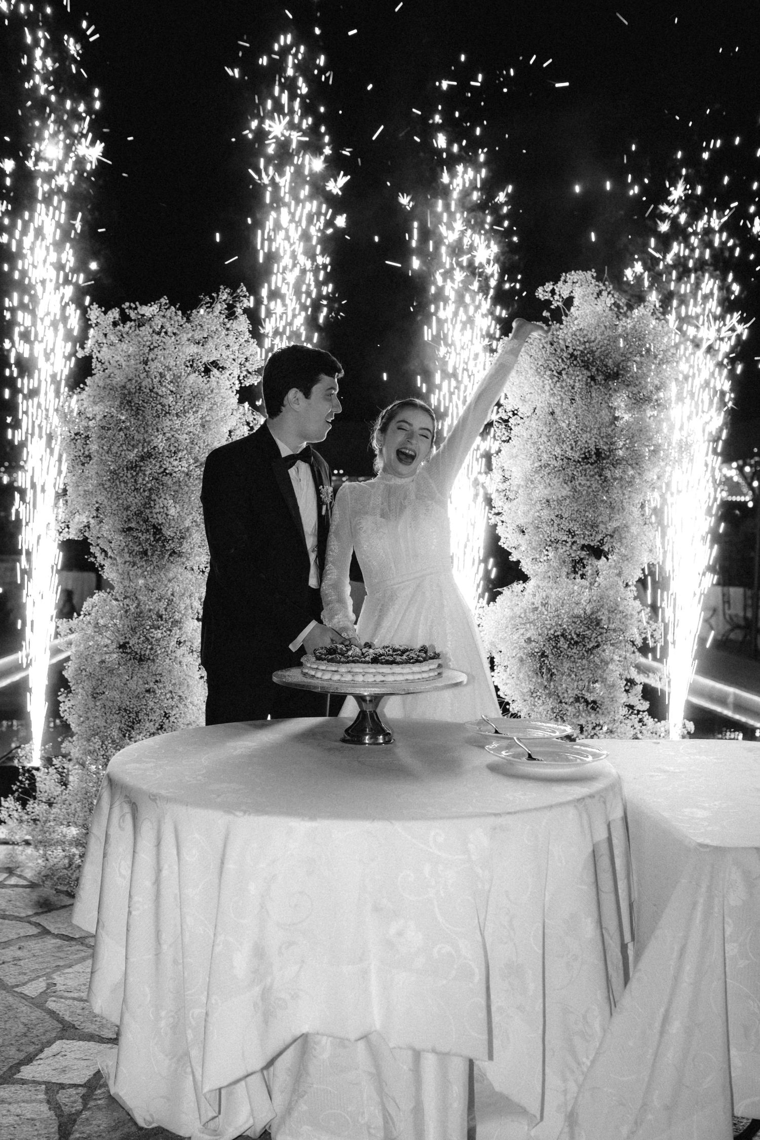 Bride and groom cutting wedding cake surrounded by spectacular fountain fireworks at night reception.