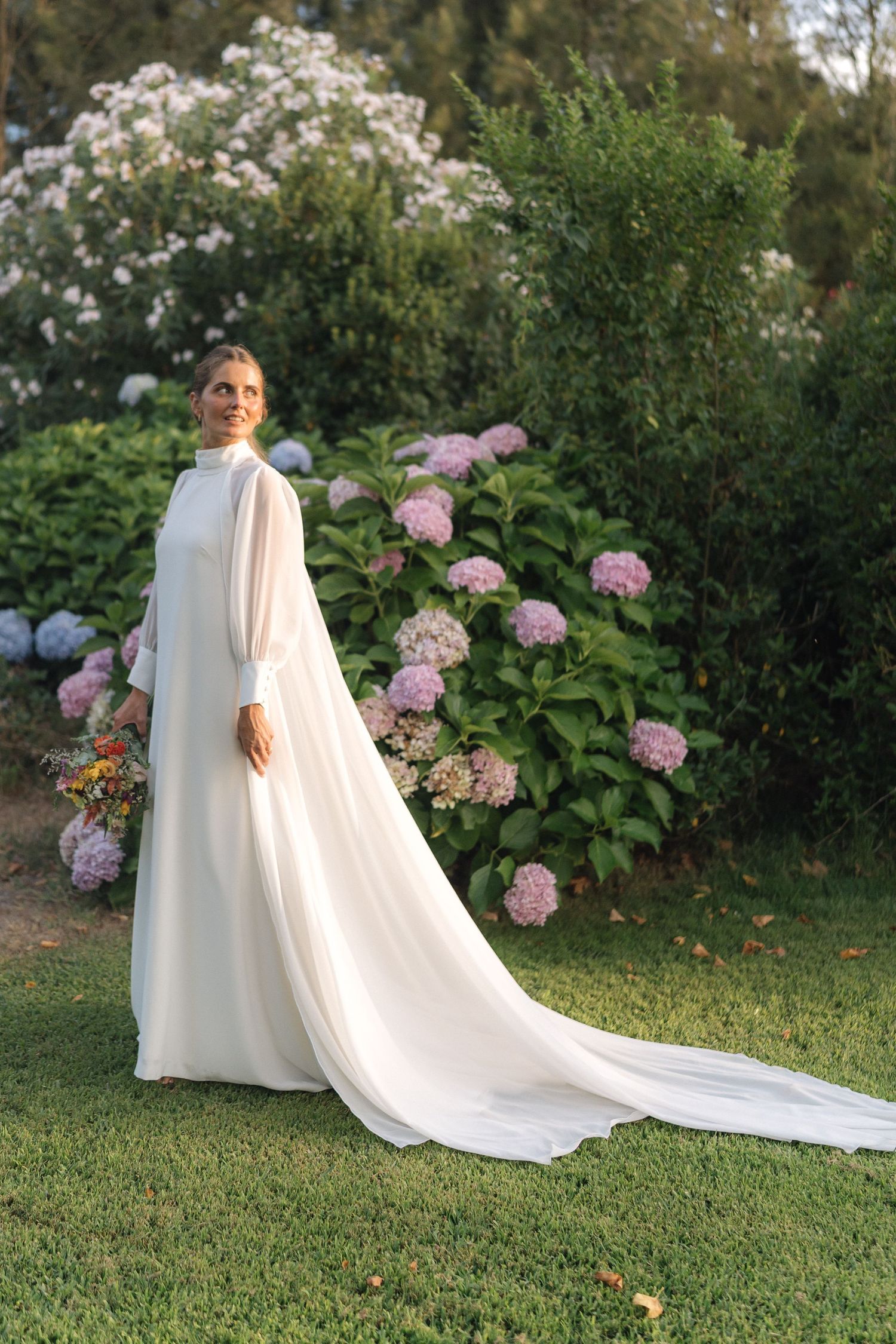 A person in a flowing white cape poses gracefully in a garden with blooming pink hydrangea flowers.