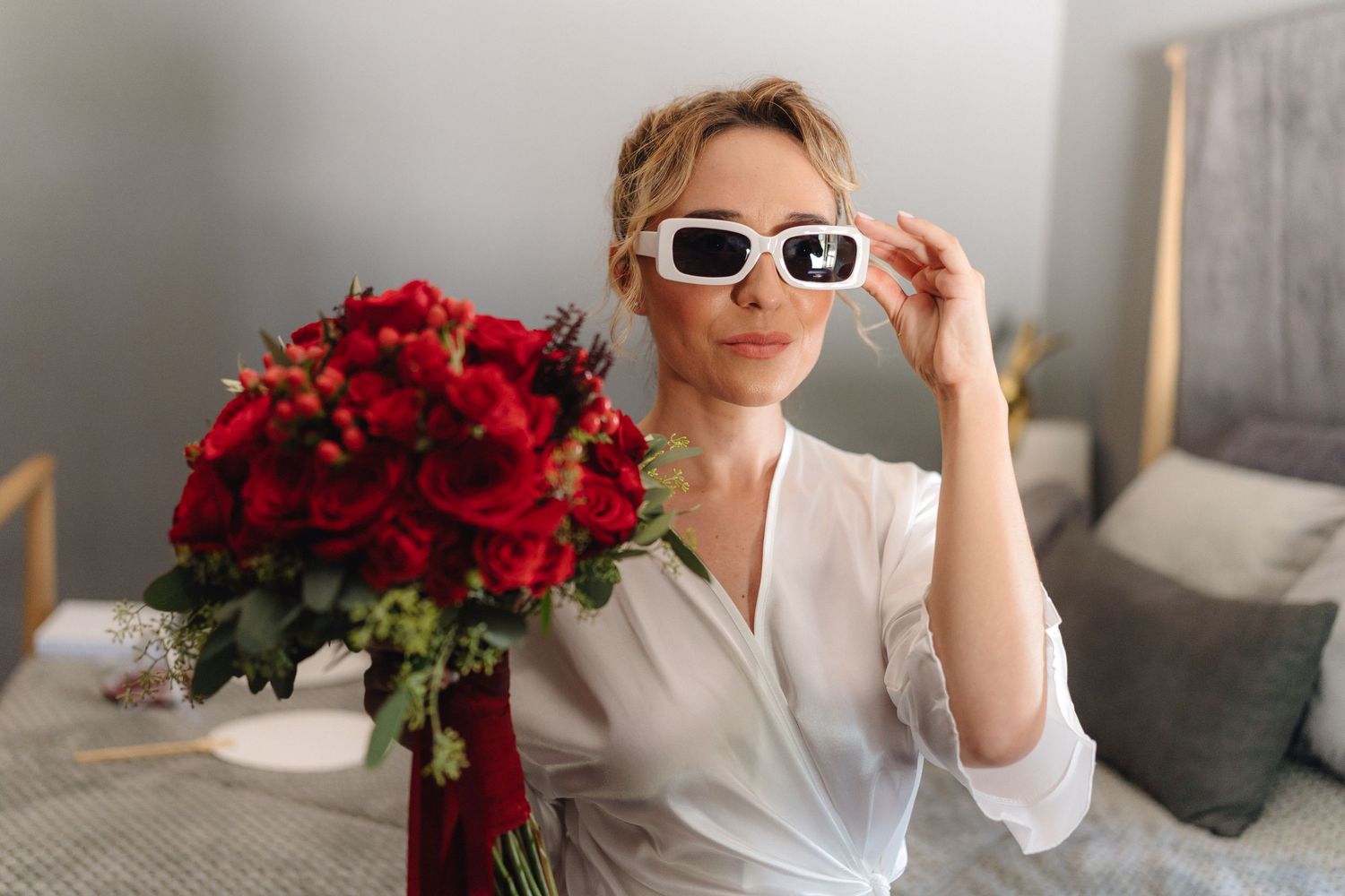 Person wearing white sunglasses poses with large red rose bouquet against grey interior background.