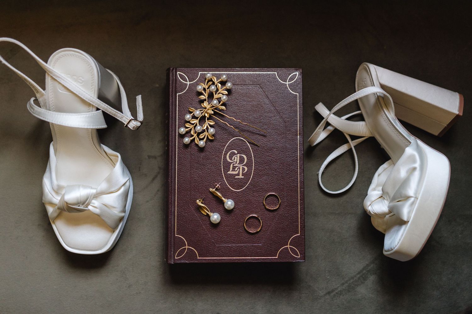 Wedding accessories including white shoes and vintage book arranged on dark surface.