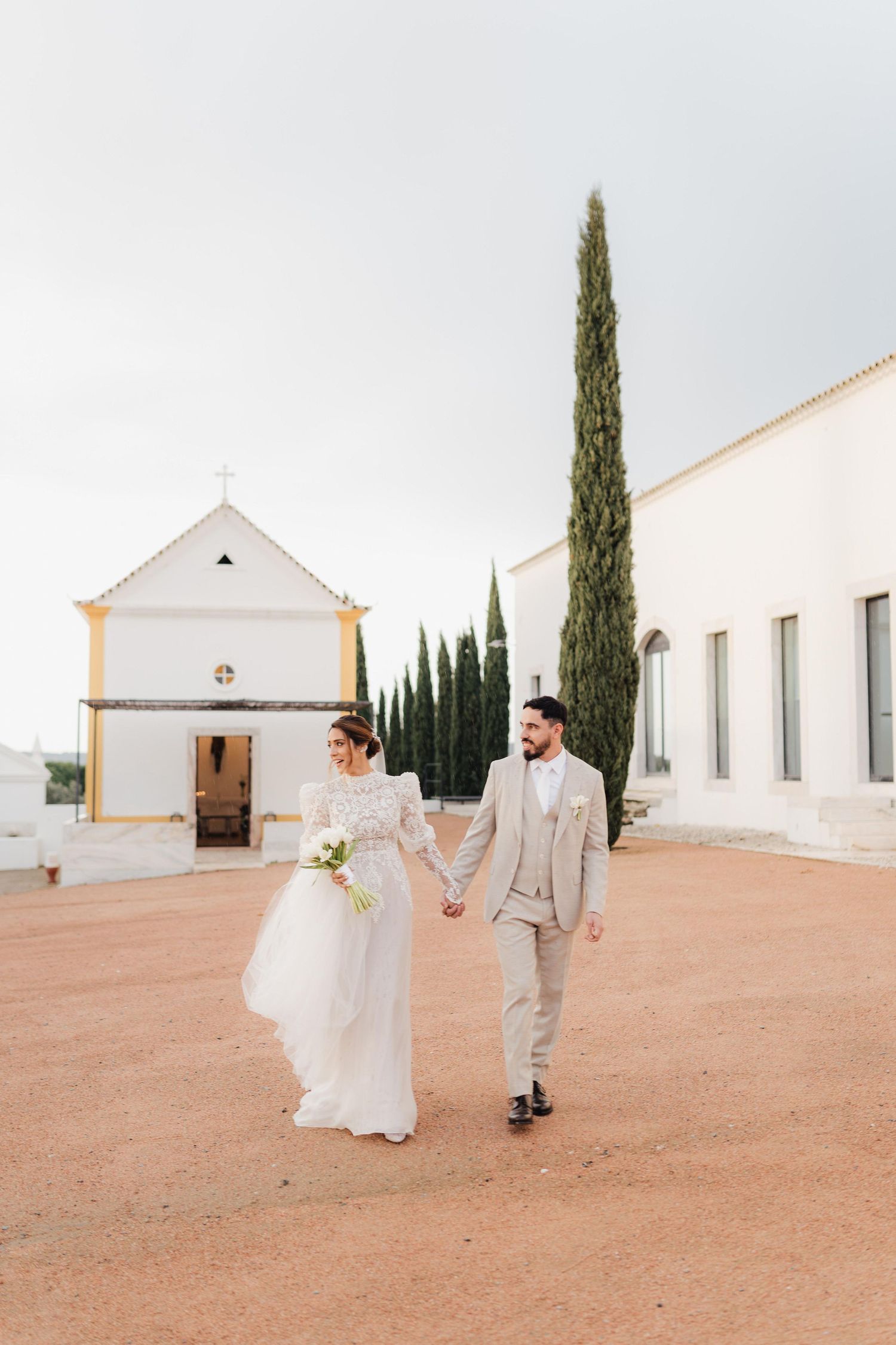 Wedding couple walking across a terracotta courtyard of a white Mediterranean-style building.