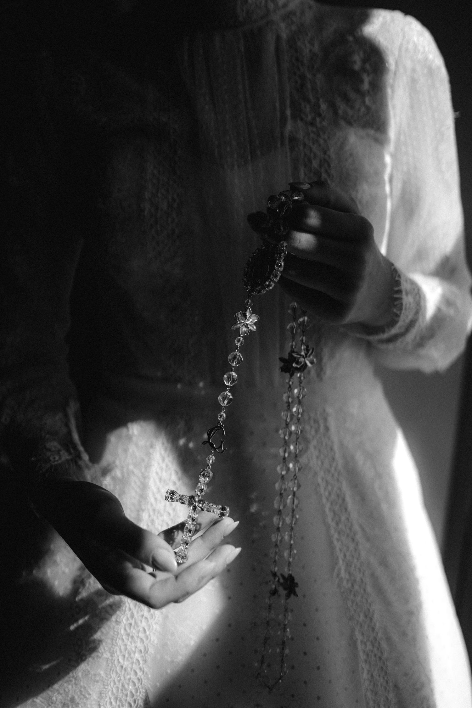 Black and white photo of a woman in lace dress holding a rosary with cross pendant in dramatic lighting.