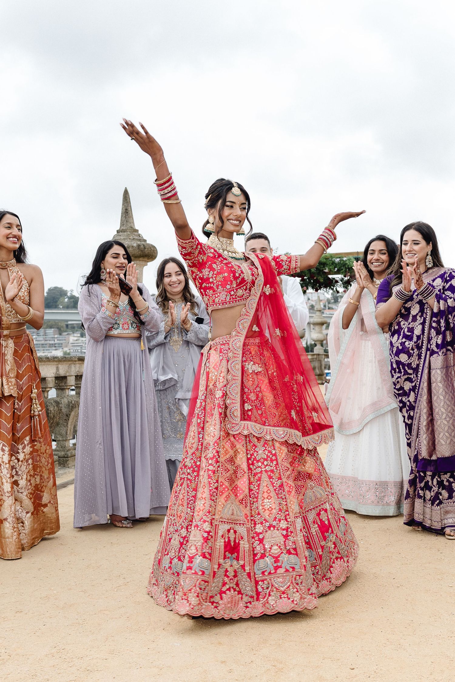 Bride in red lehenga dances joyfully surrounded by guests in colorful formal attire.
