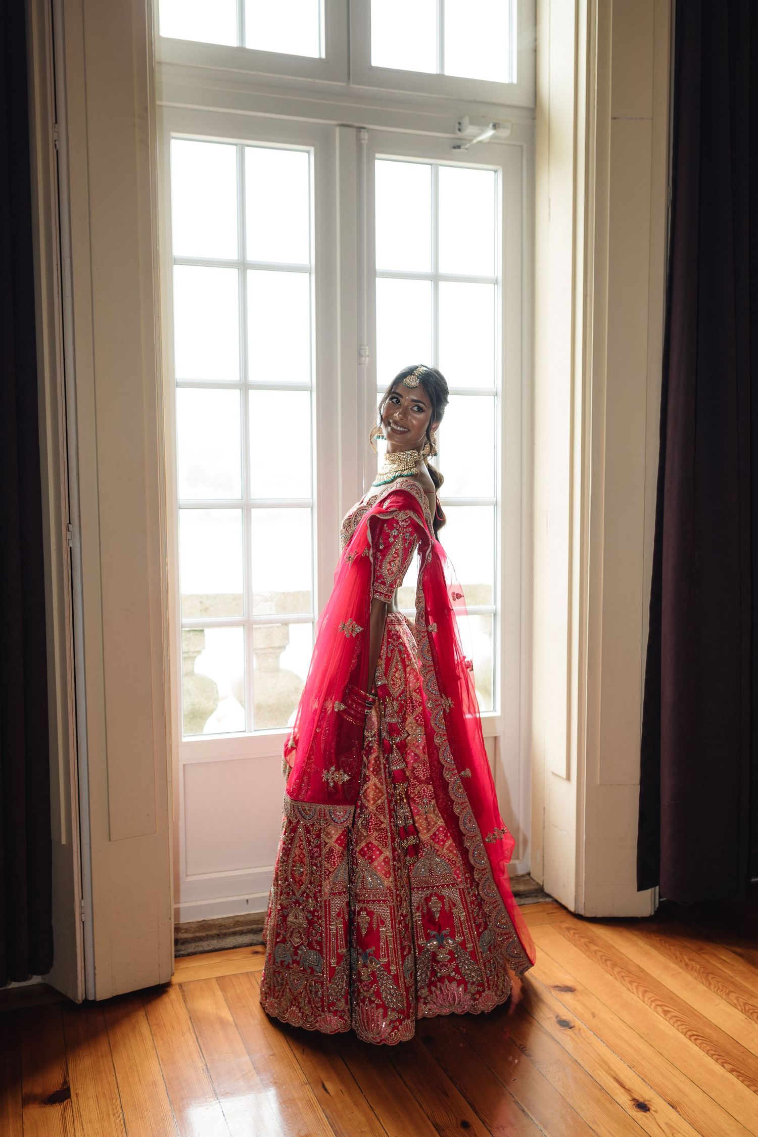 Ornate red and gold wedding lehenga showcased in sunlit room with hardwood floors.