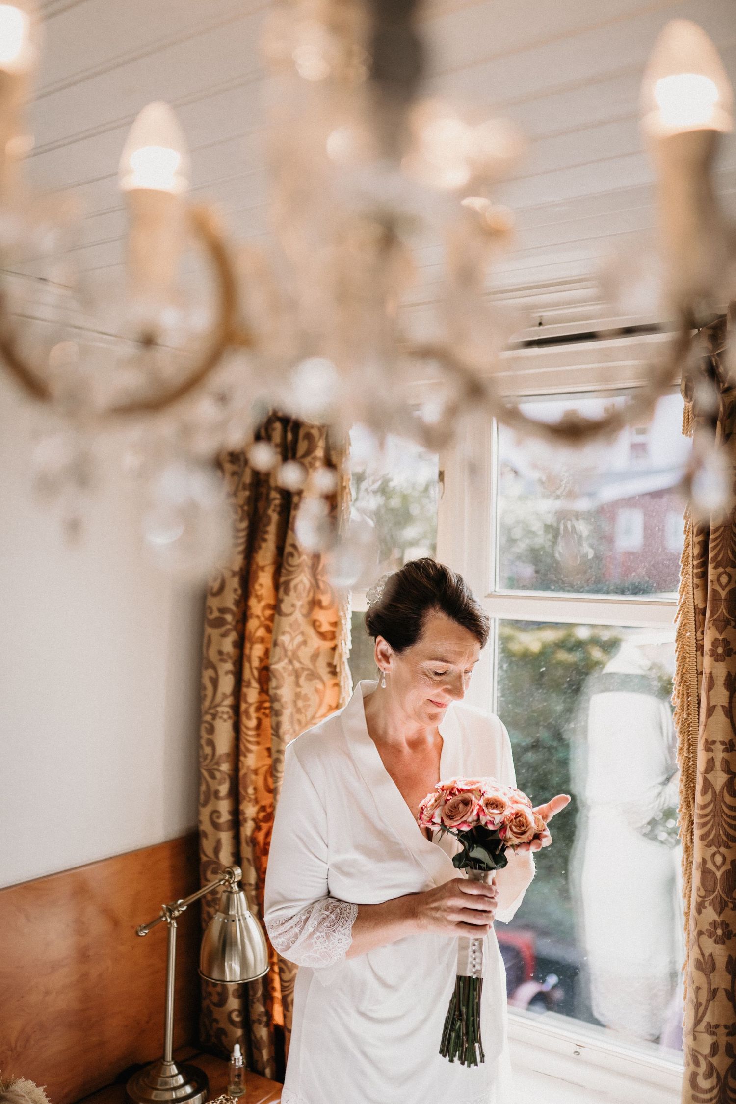 A person in white clothing holds and smells a bouquet near a chandelier.