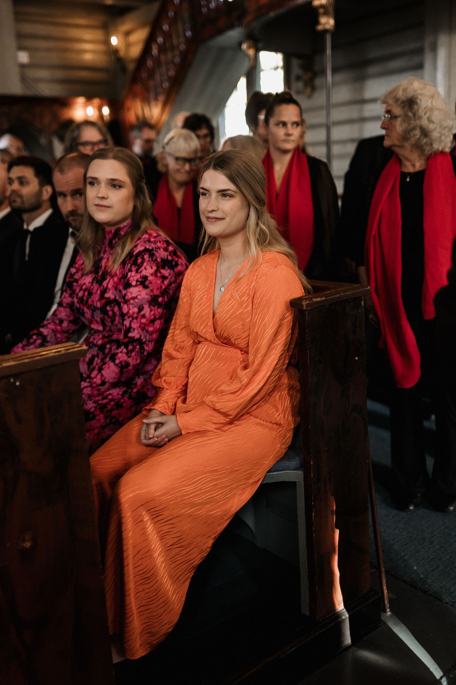 Person in bright orange dress seated among crowd wearing red in ornate venue.