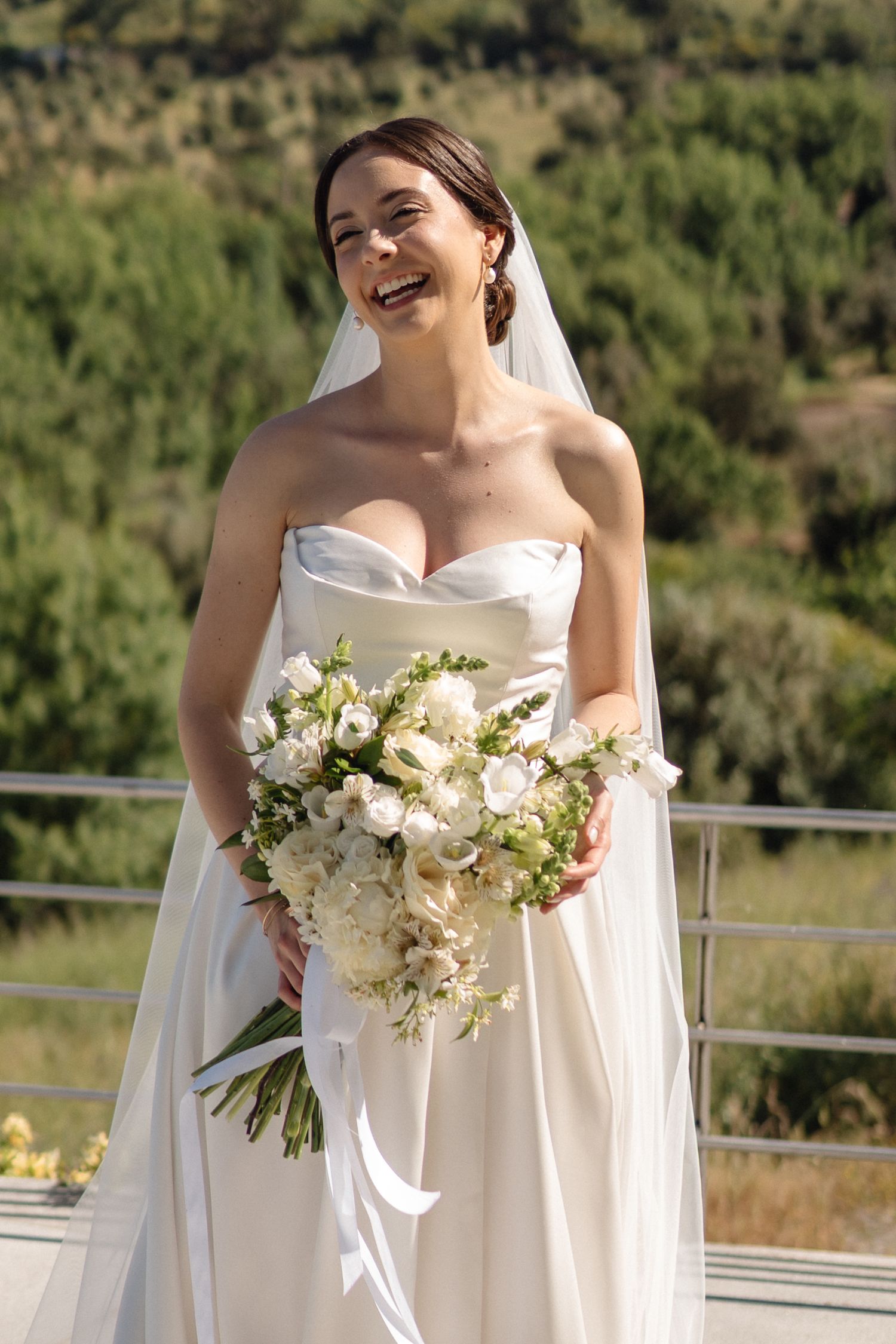 A radiant bride in a strapless white gown laughs joyfully while holding a lush white and cream bouquet outdoors.