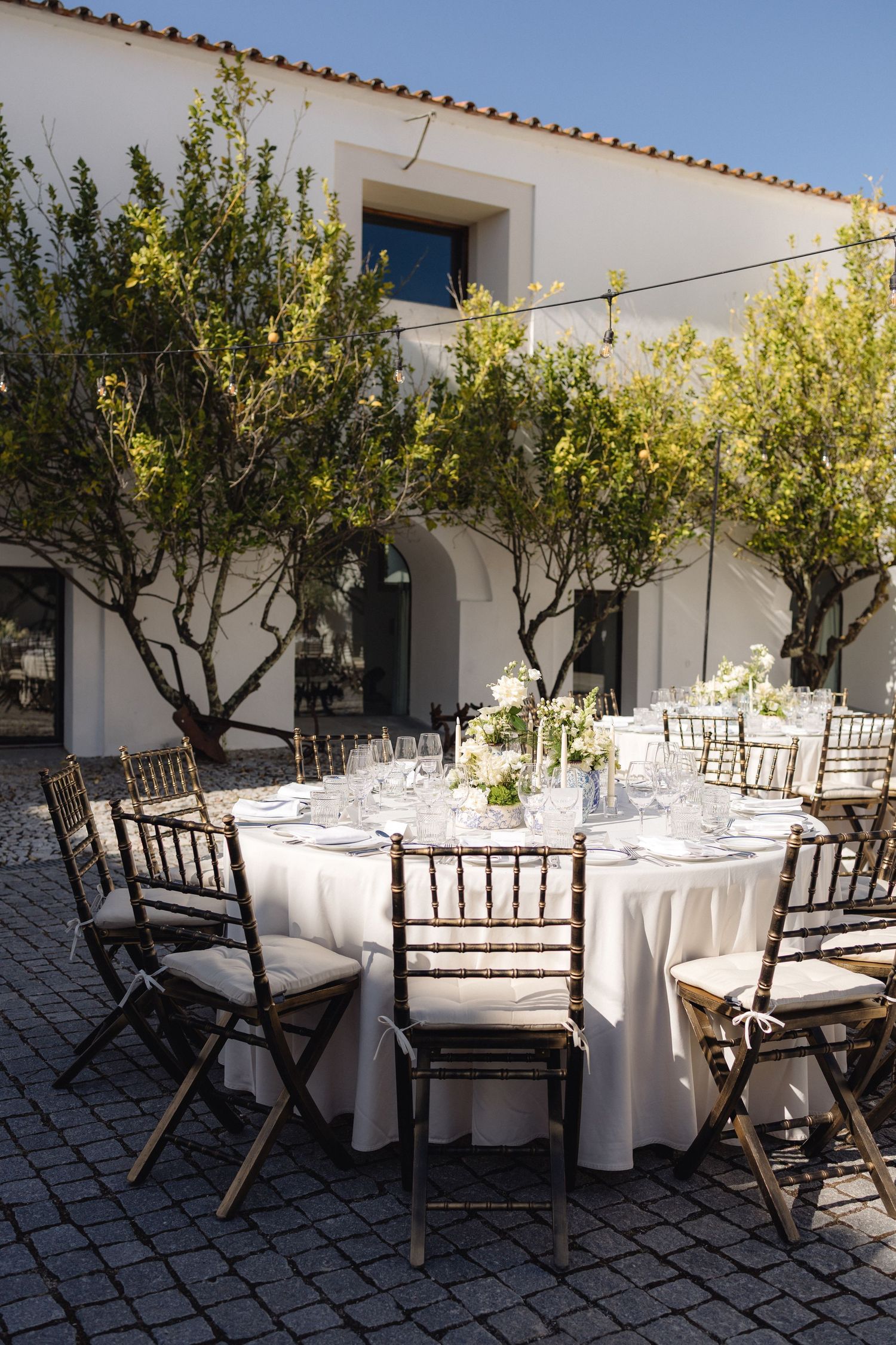 Outdoor dining area with round tables and wooden chairs arranged under olive trees in a stone courtyard.