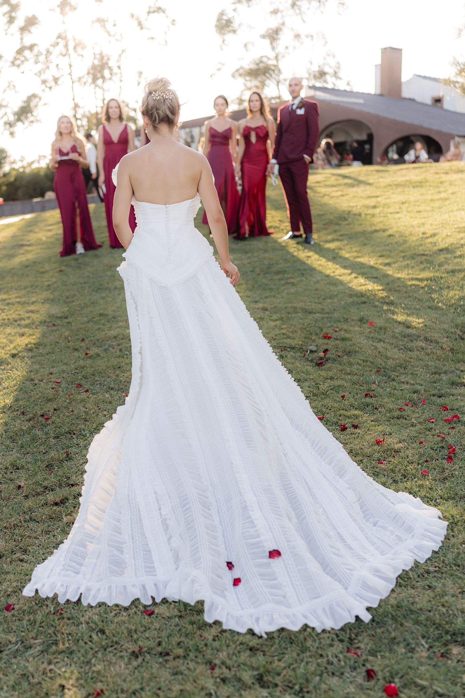 White lace wedding gown with long train displayed on green lawn with scattered rose petals.