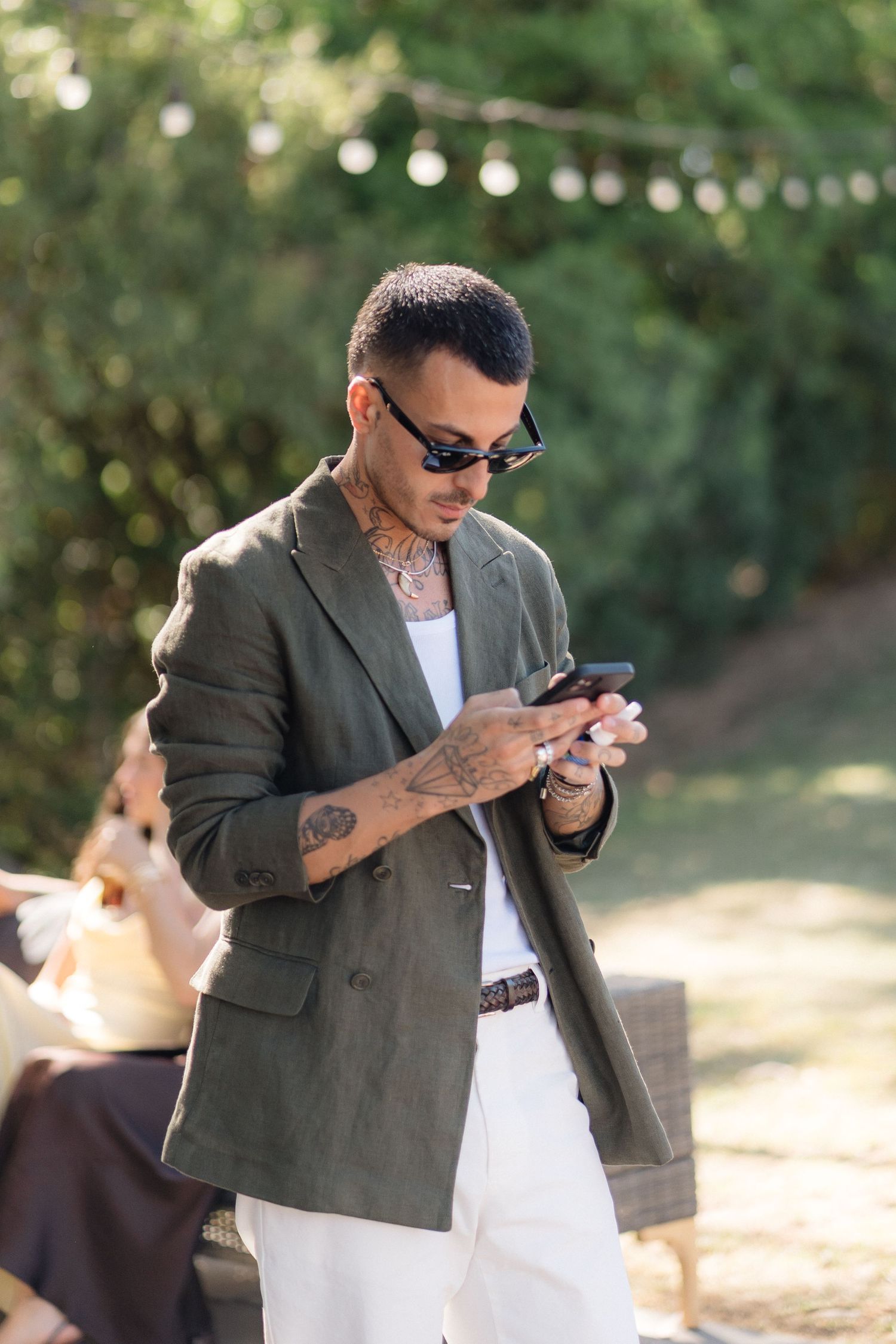 Stylish guest in grey blazer and sunglasses checks phone at outdoor event.