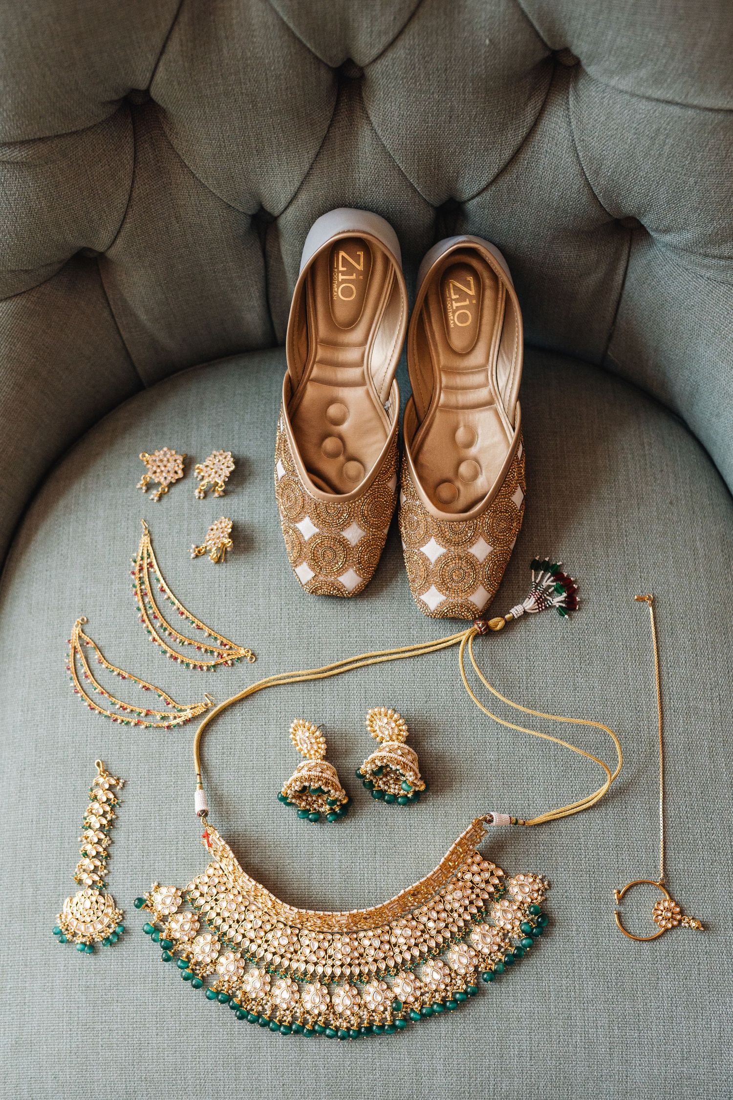 Gold embellished bridal shoes and traditional Indian jewelry set on grey tufted chair.