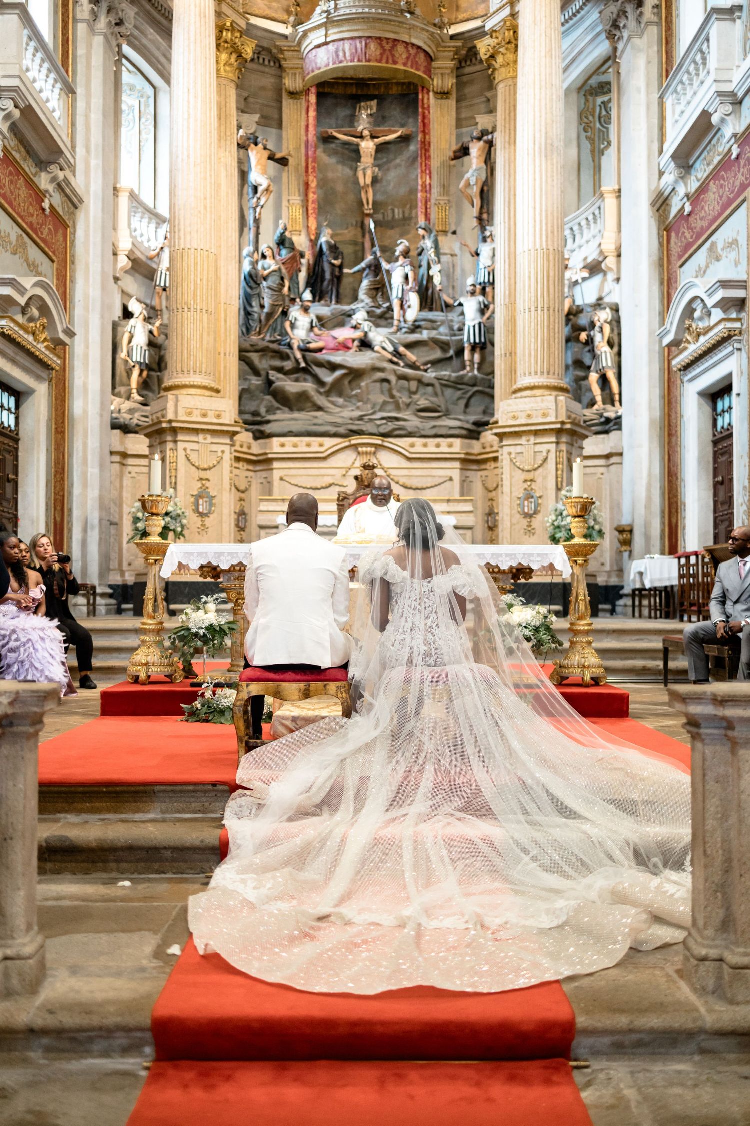 A wedding ceremony in an ornate cathedral with a long flowing veil and red carpet leading to an elaborate altar.