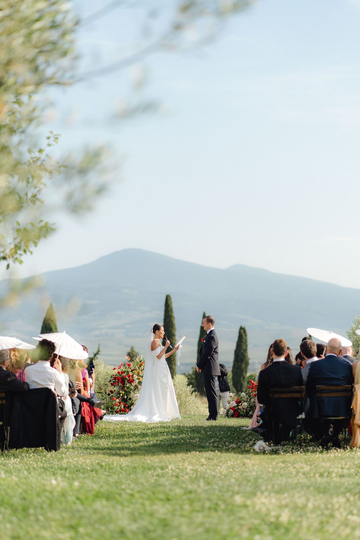Outdoor wedding ceremony on a green lawn with Tuscan hills and cypress trees in the background under a clear blue sky.