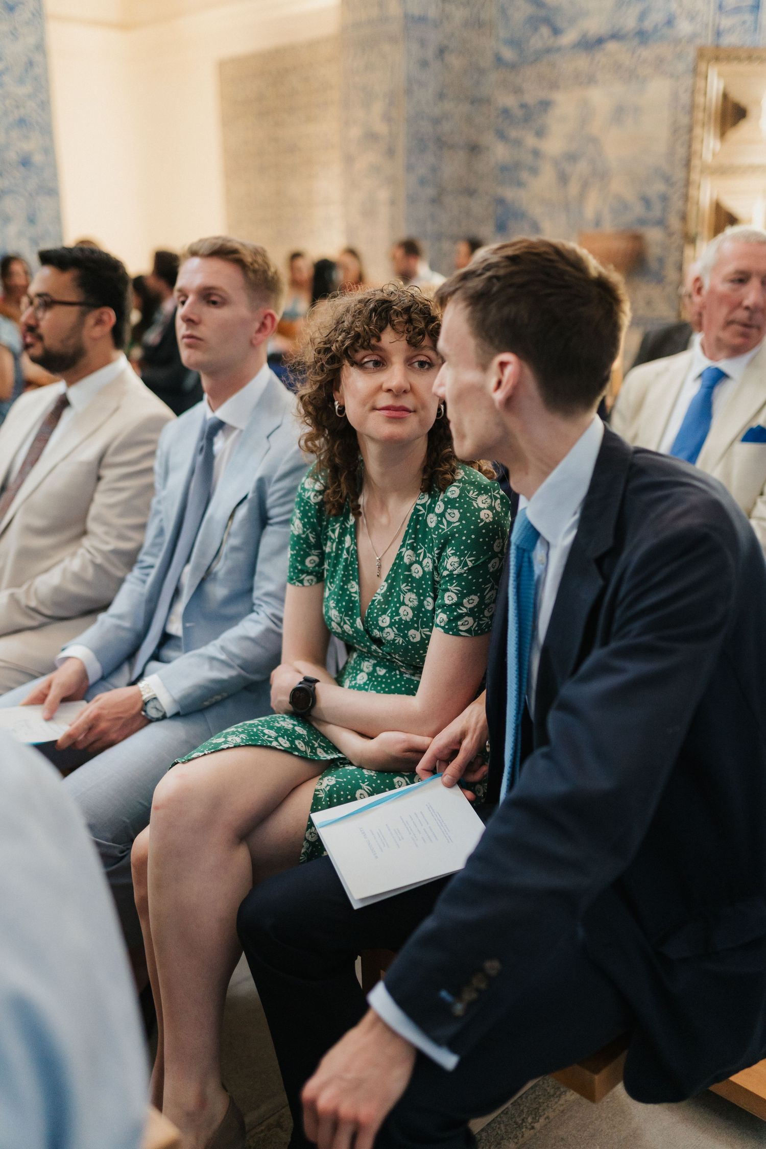 Wedding guests in formal attire seated in rows during an indoor ceremony.
