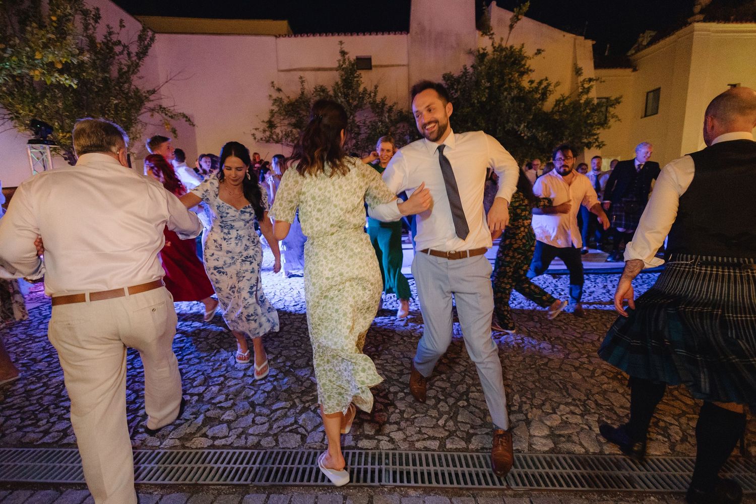 Wedding guests dancing together at an outdoor evening reception with blue lighting and cobblestone ground.