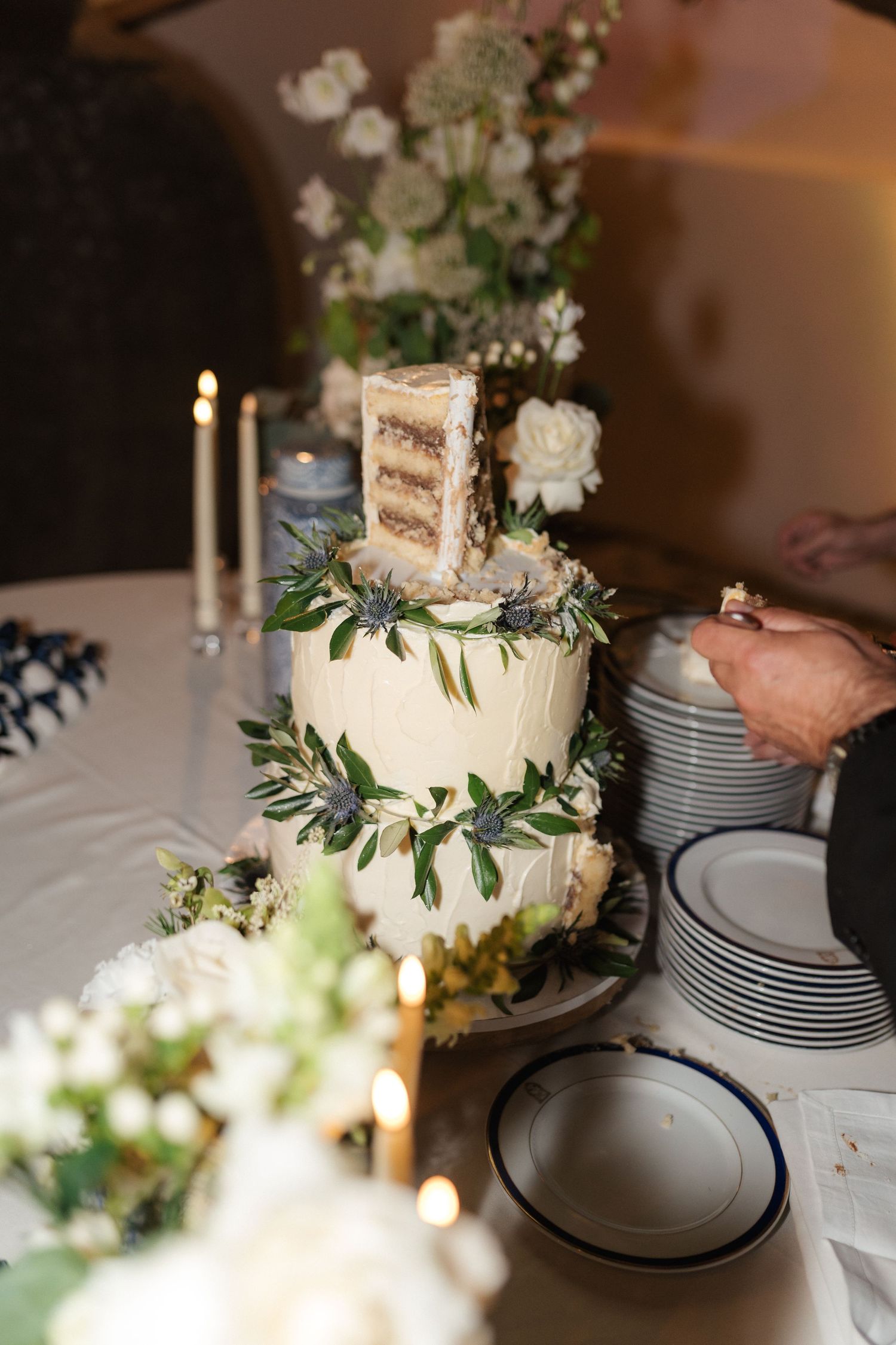 Elegant white wedding cake with greenery decorations displayed next to stacked plates for serving.