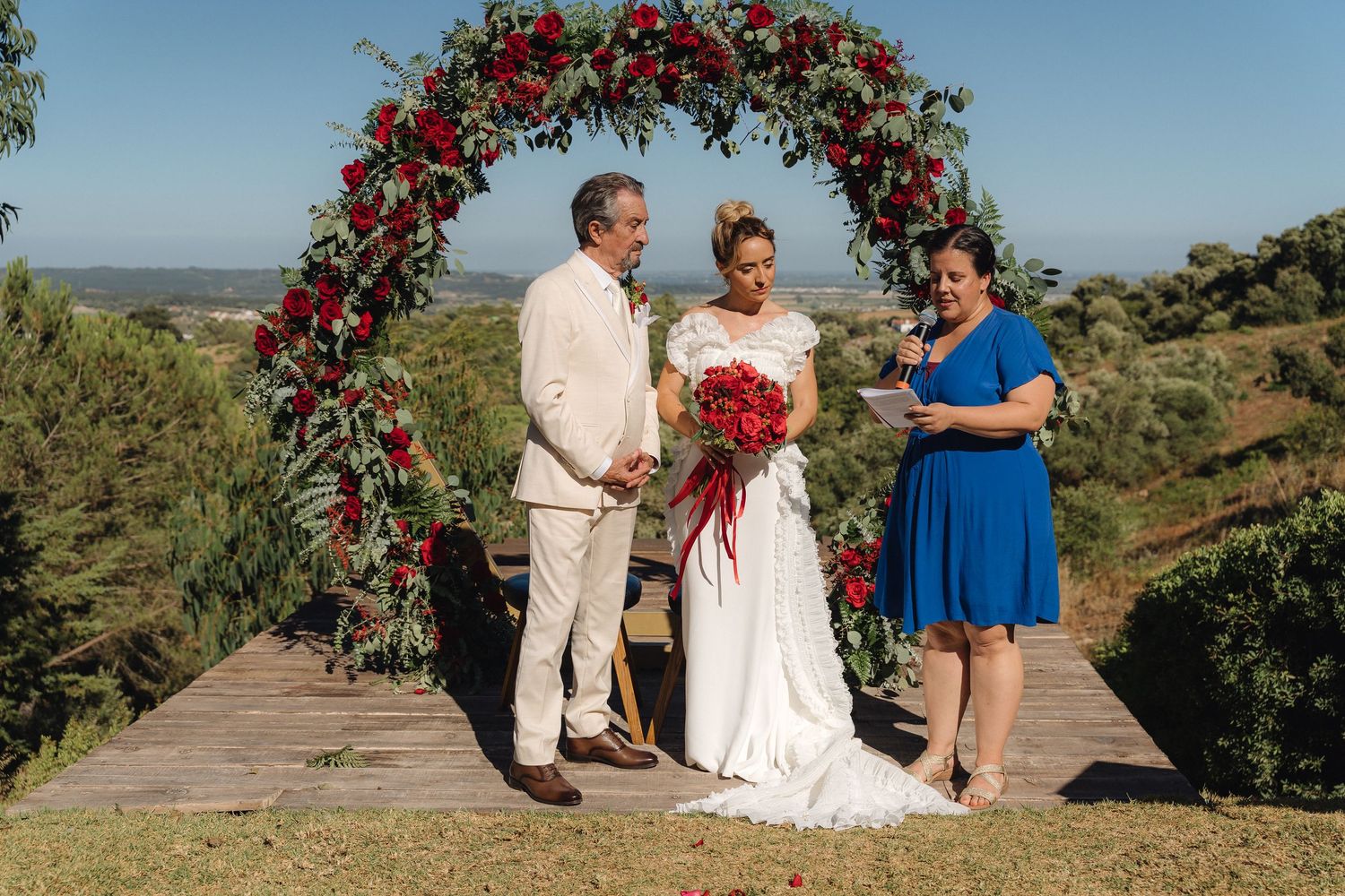 Wedding ceremony participants gather under a stunning red rose archway in a hilltop setting.