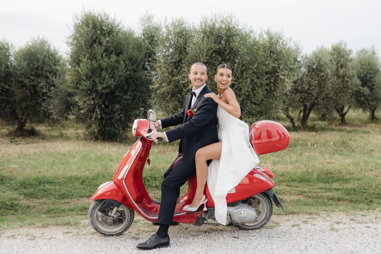 Smiling bride and groom sitting together on a red Vespa scooter surrounded by olive trees.