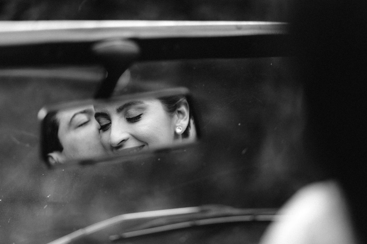 Black and white photo of a couple sharing a romantic moment reflected in a car rearview mirror.