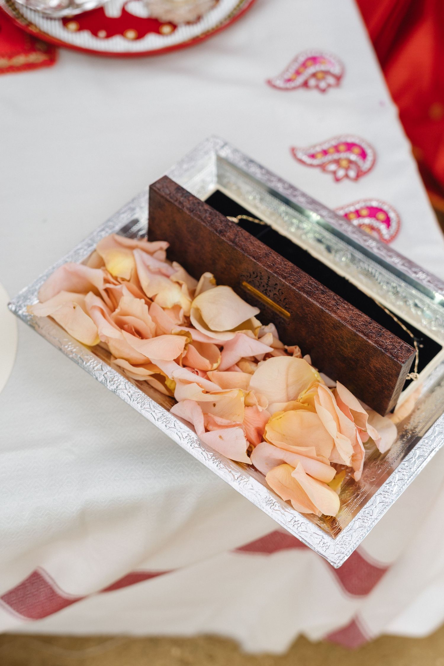 Decorative silver tray with rose petals and wooden box on white and red patterned fabric.