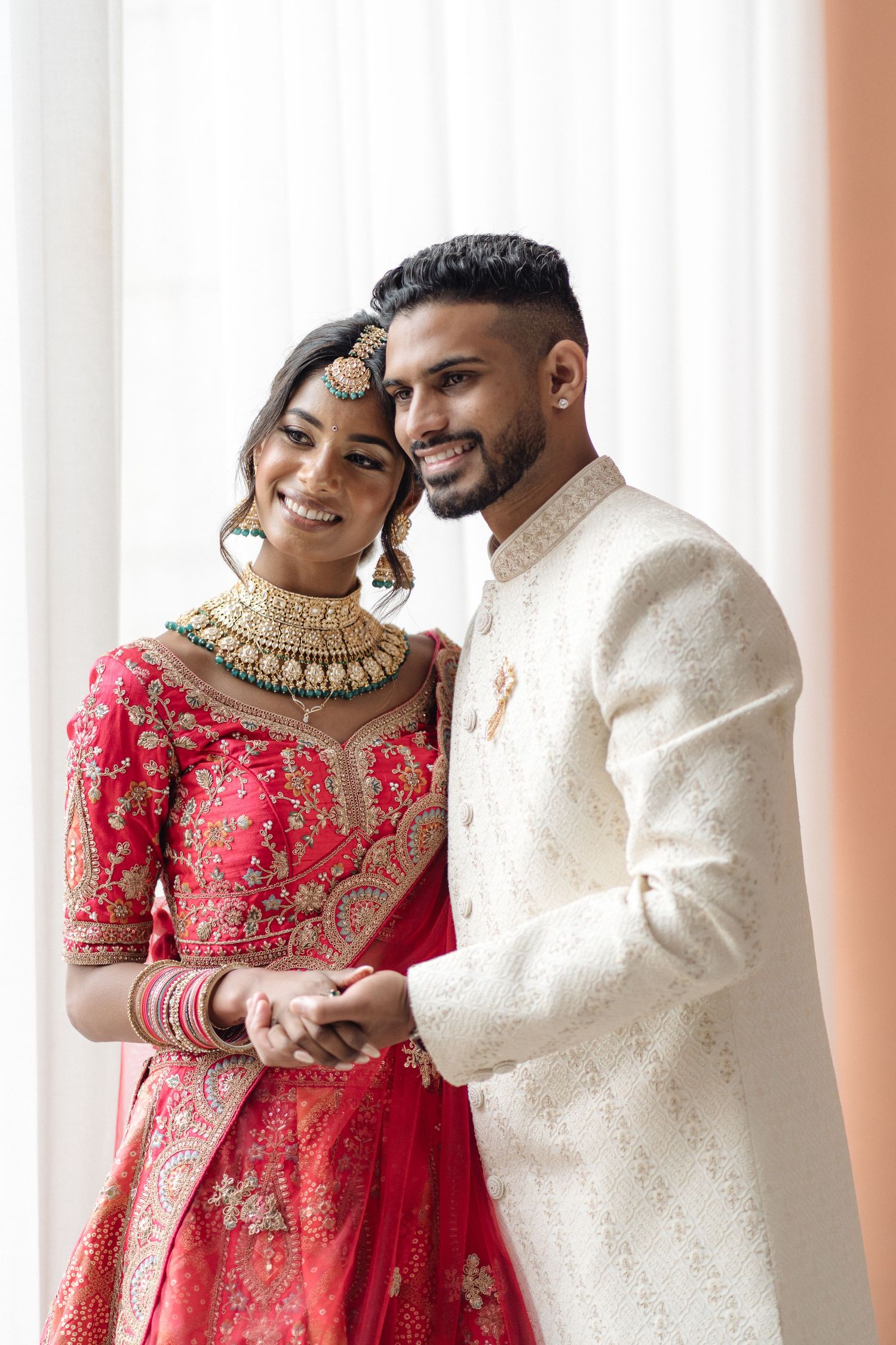 A couple in traditional South Asian wedding attire, with the bride in red lehenga and groom in white sherwani embracing.