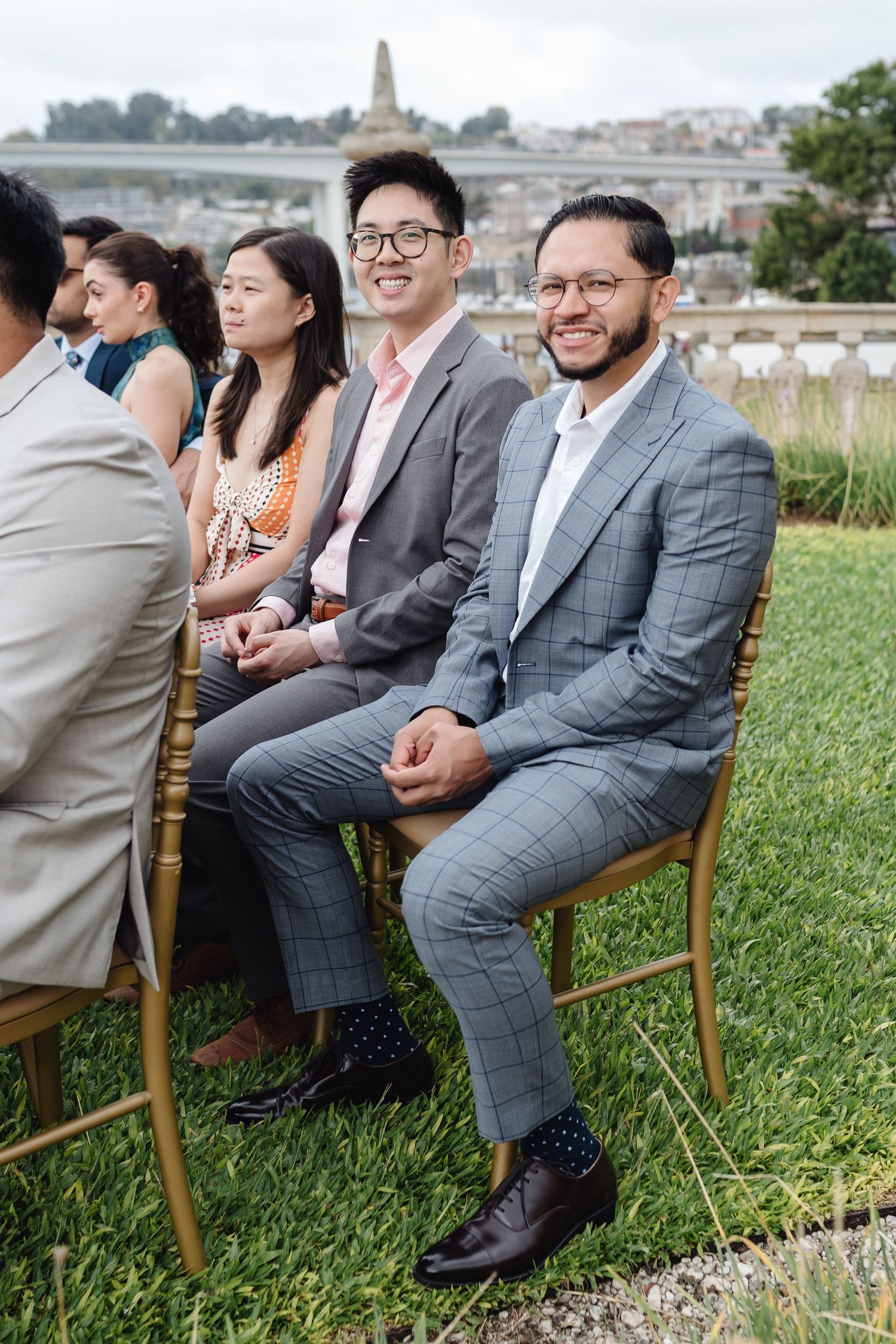Wedding guests in formal attire seated on wooden chairs on a grassy lawn with city views.