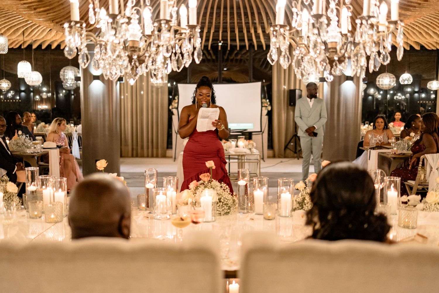 Person in red dress gives a speech at an elegant wedding reception with crystal chandeliers.