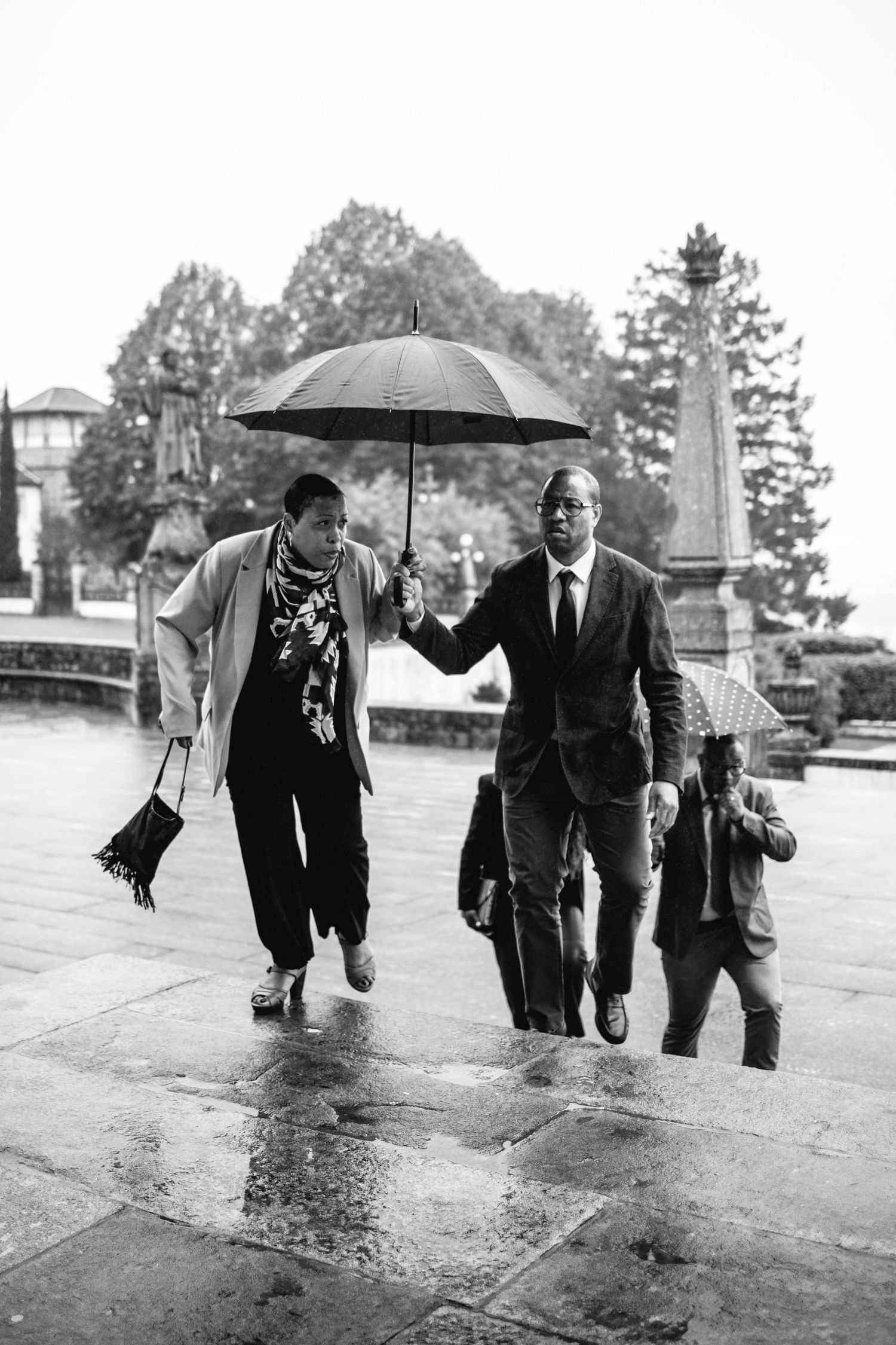 A black and white photo of wedding guests walking under an umbrella in the rain.