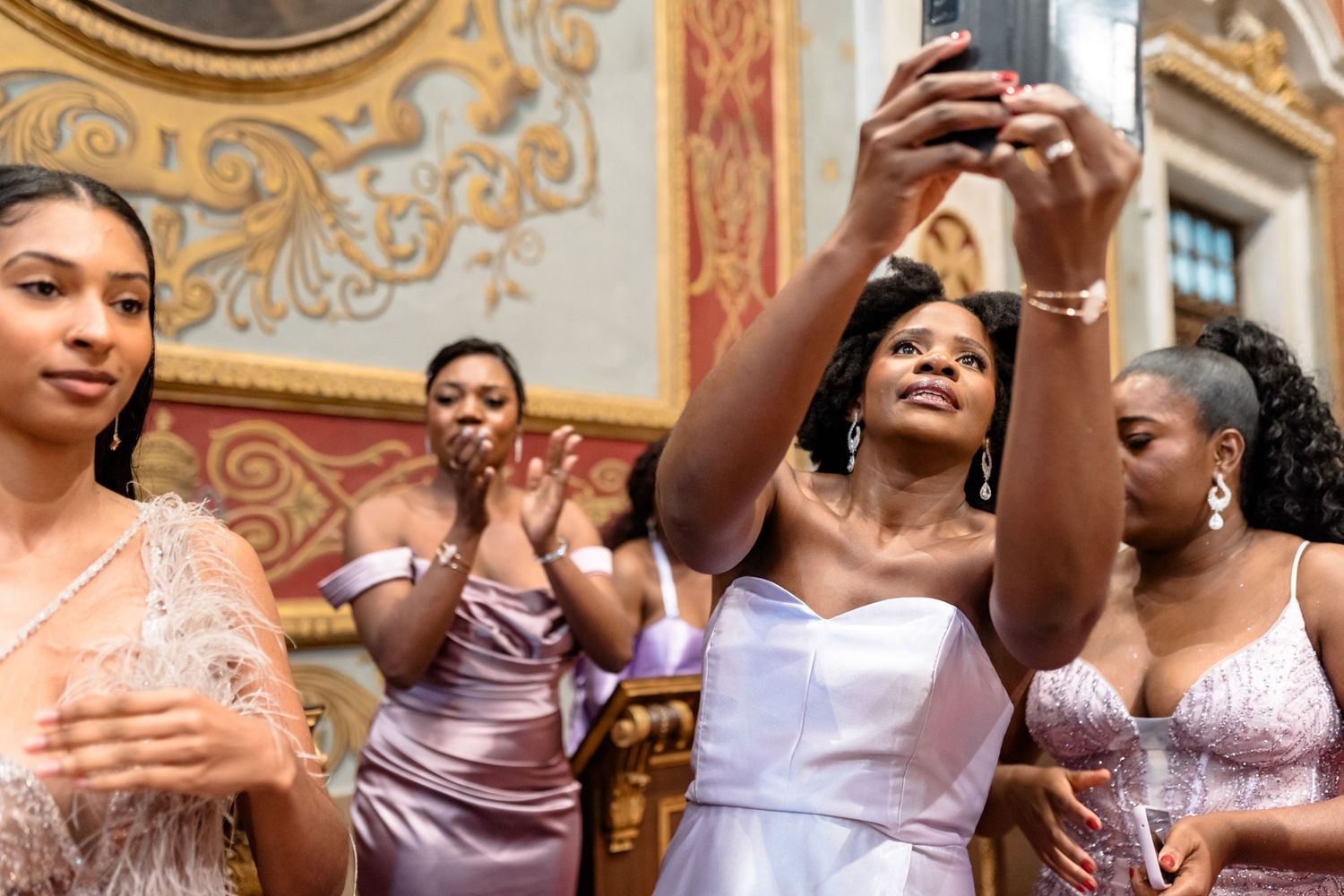 Wedding party members pose together in elegant purple and white attire at an ornate venue.