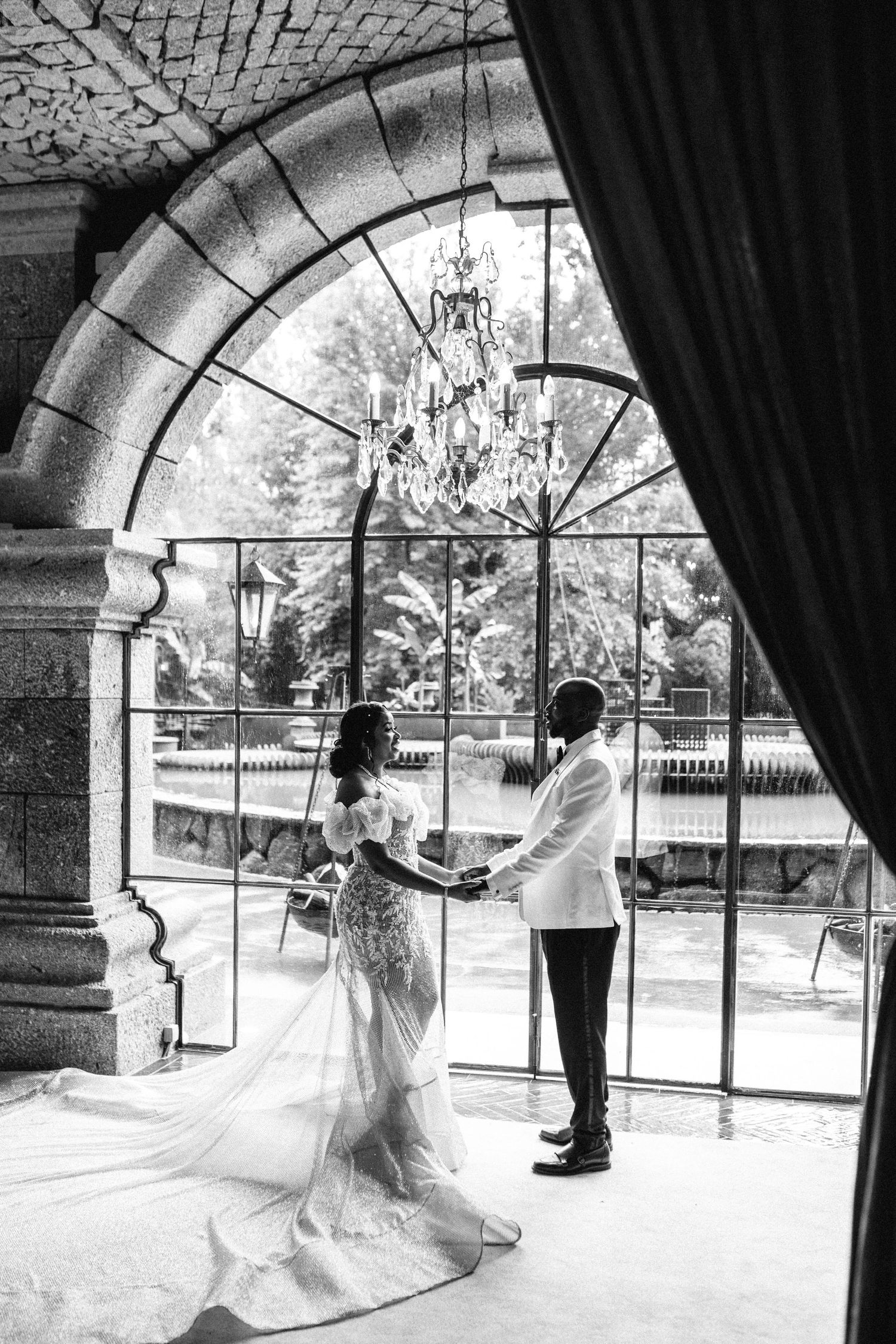 Wedding couple dancing in an elegant arched room with chandelier and natural lighting.