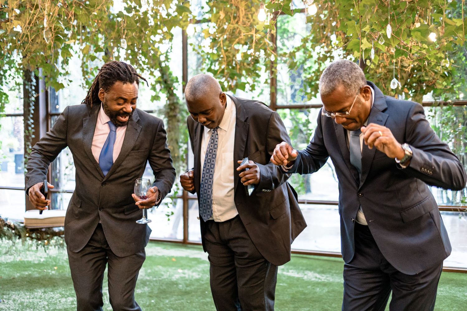 Three groomsmen in dark suits dance and celebrate together at an outdoor wedding venue.