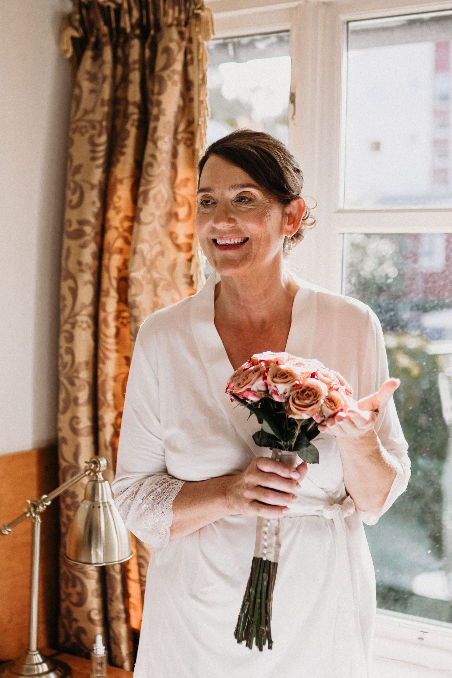 Person in white holding a bouquet of pink roses near a window with golden curtains.