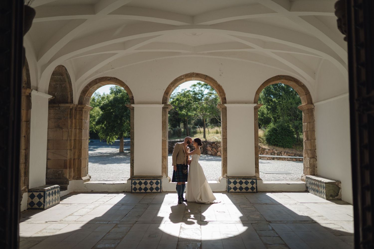 A sunlit stone colonnade with arched windows creates a dramatic setting for wedding photography.