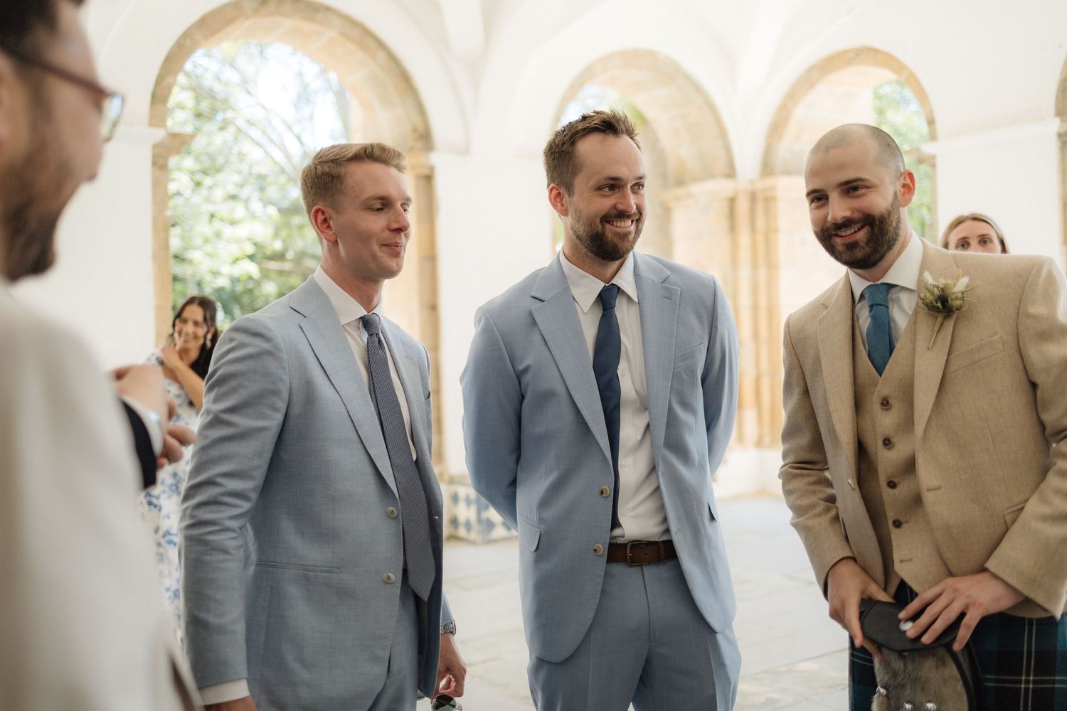 Groomsmen in light gray and tan suits gather in bright hallway.