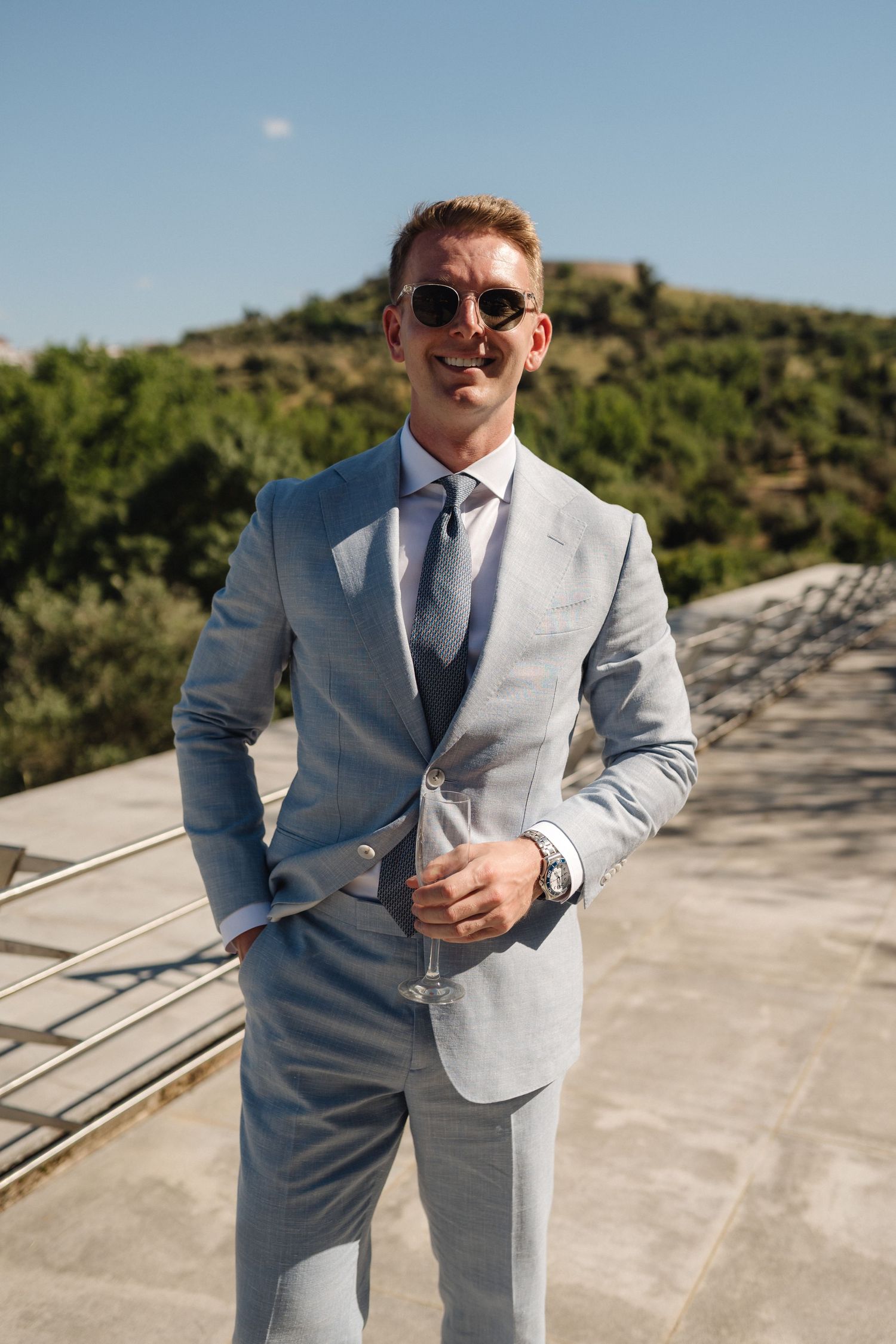 Stylish groomsman in a light grey suit poses outdoors on a sunny day.