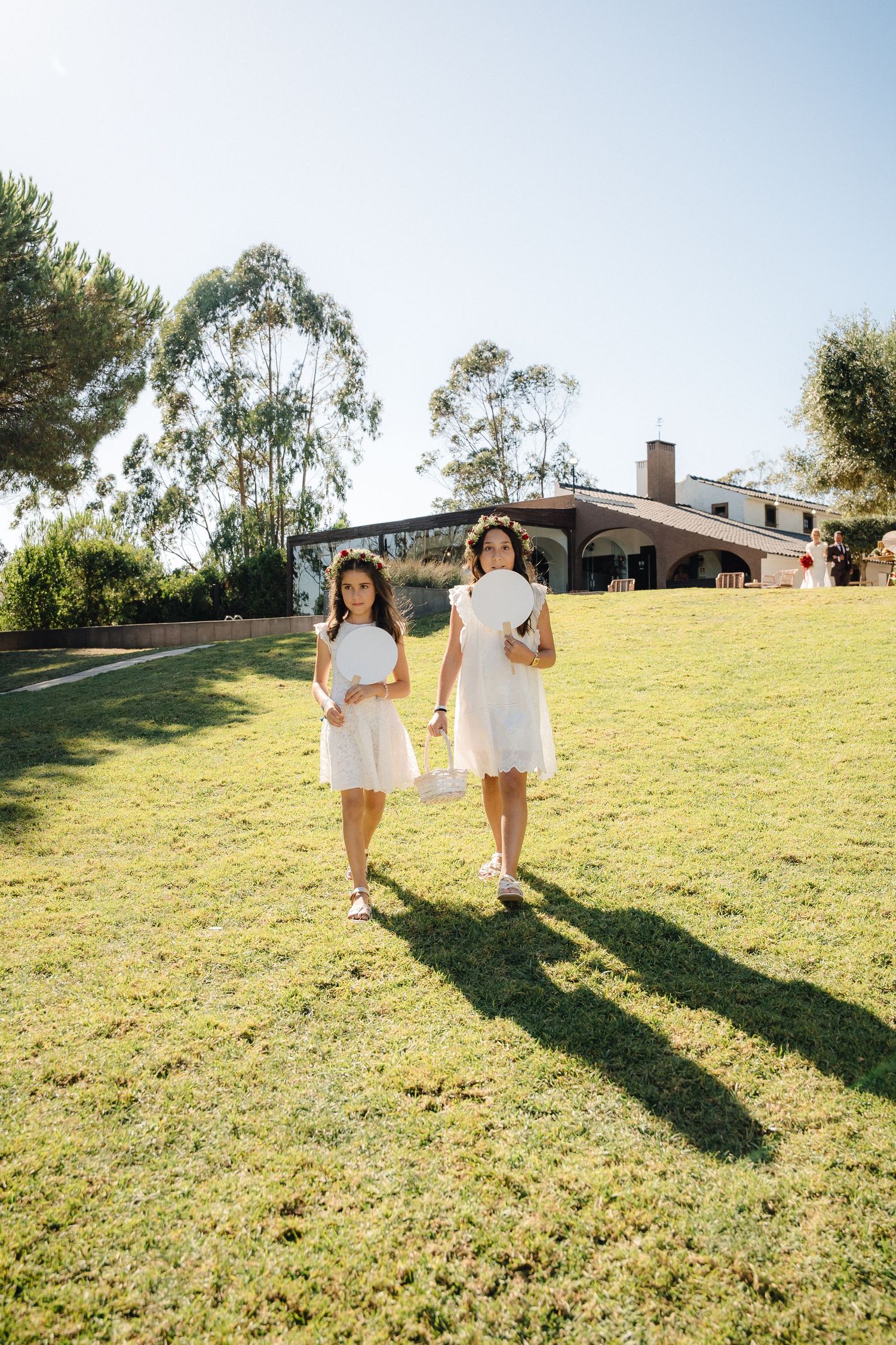 Two people in white dresses walk across a sunny lawn carrying flower baskets.