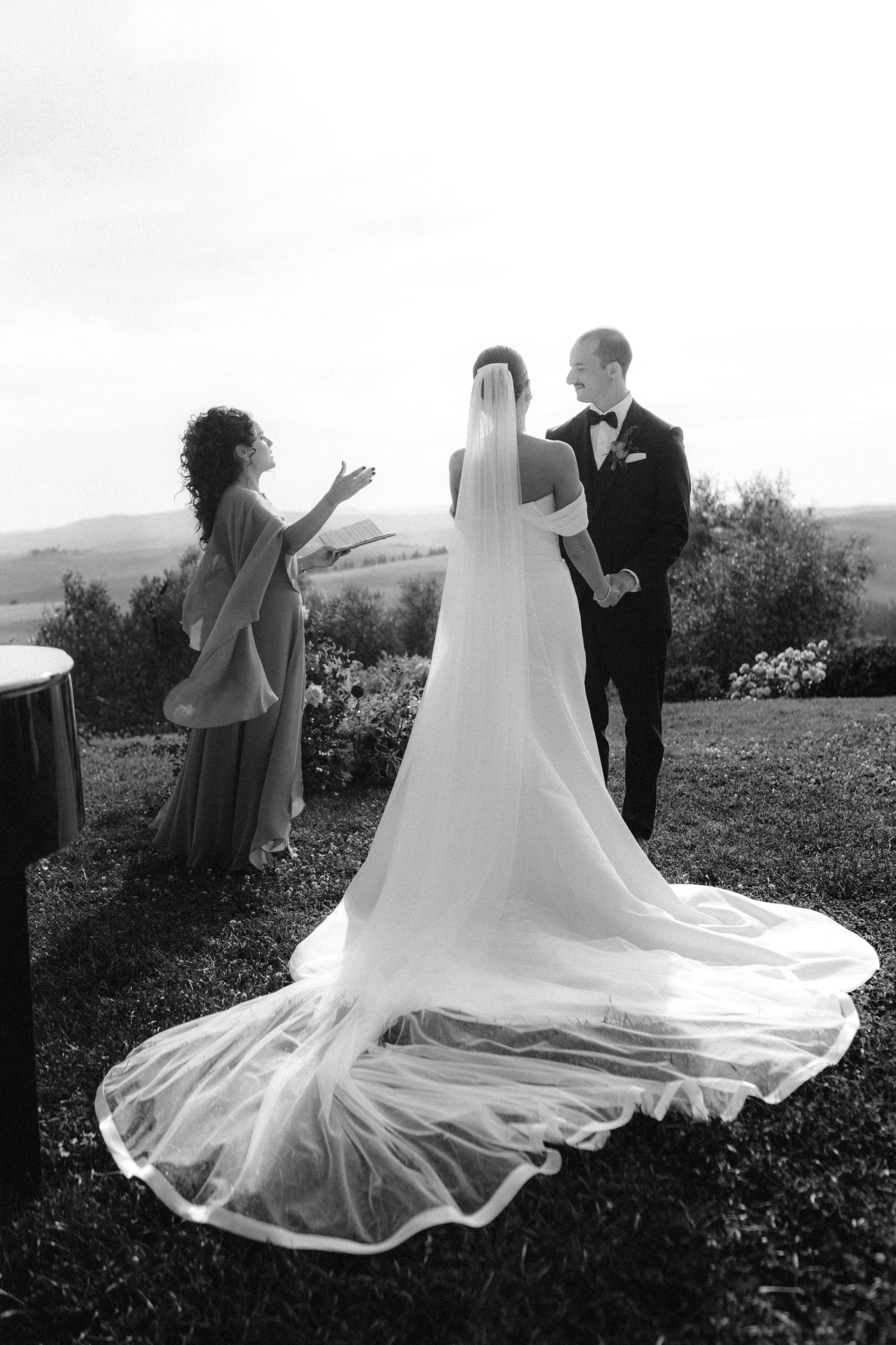 Black and white photo of bride in flowing veil and groom in tuxedo at outdoor wedding ceremony.