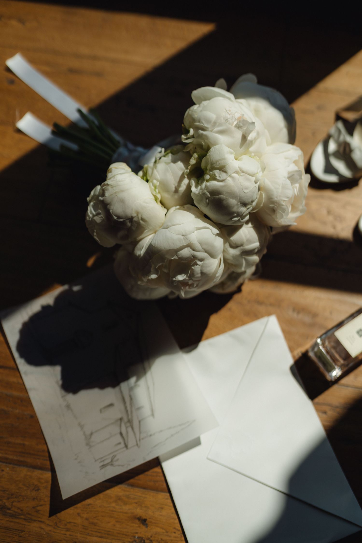 White peony bouquet resting on wedding stationery and envelope on a wooden table in warm sunlight.