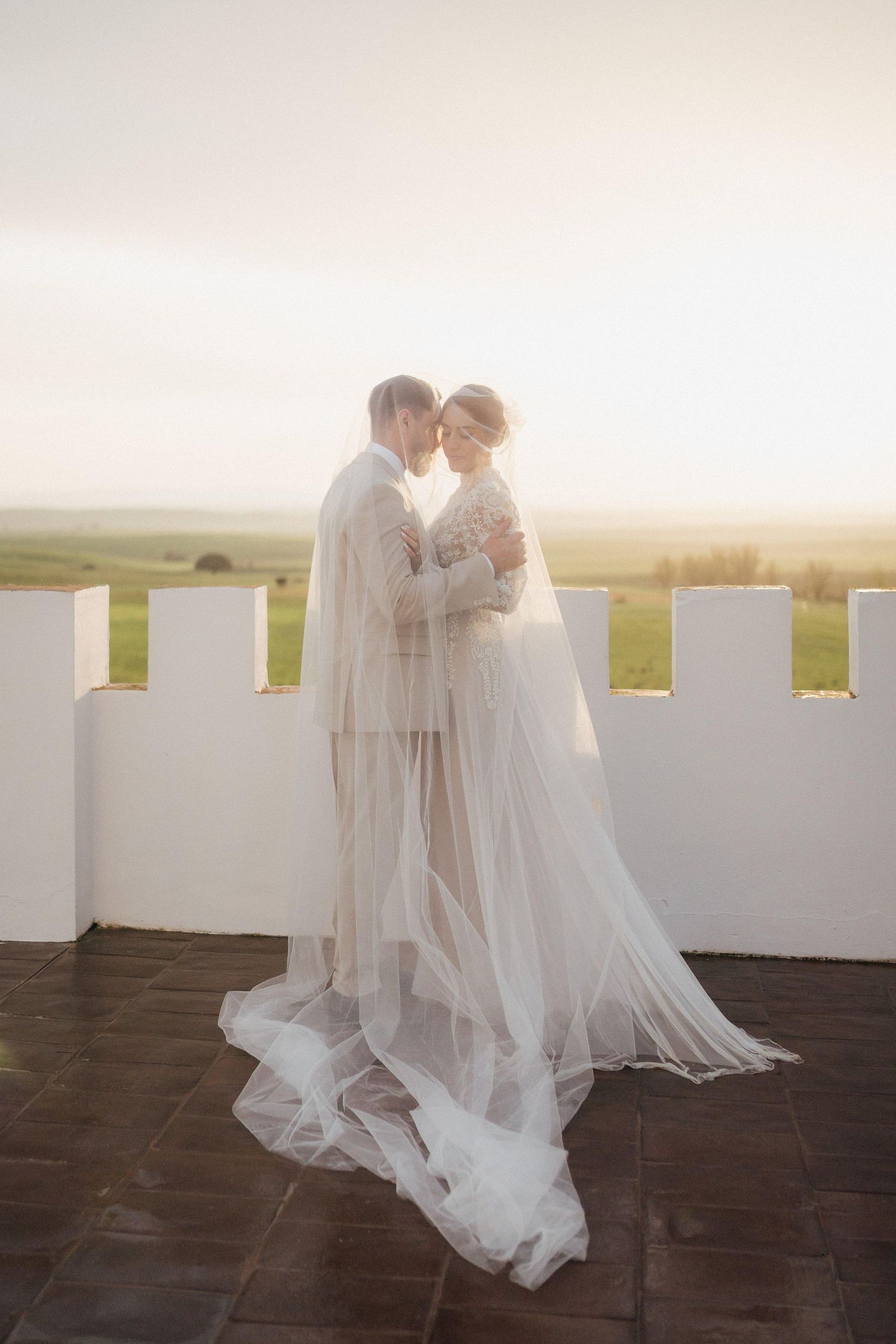 Newlyweds share an intimate moment on a castle rooftop terrace at sunset with flowing white dress and veil.