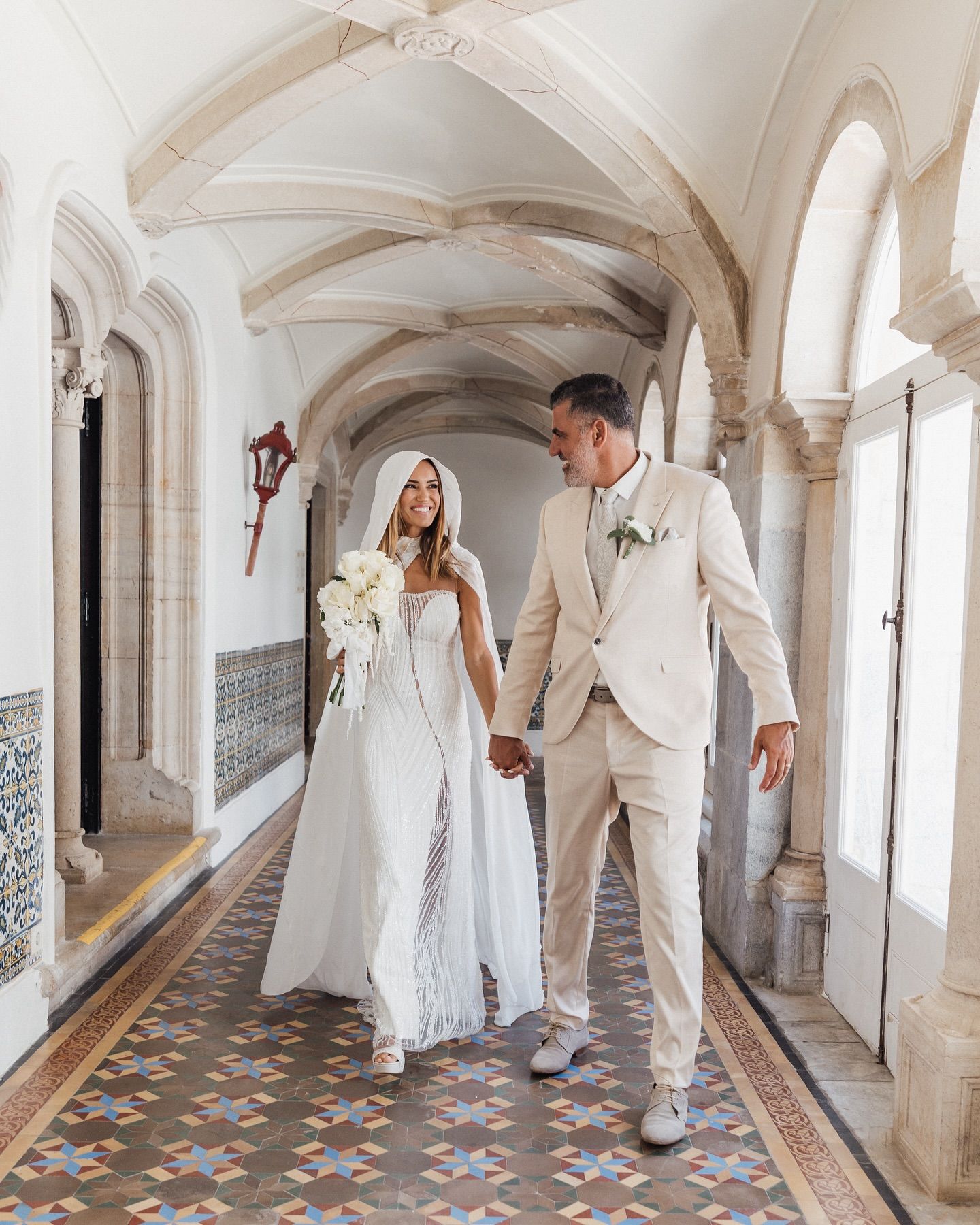 Wedding couple in white attire walking through an ornate corridor with patterned floor tiles.