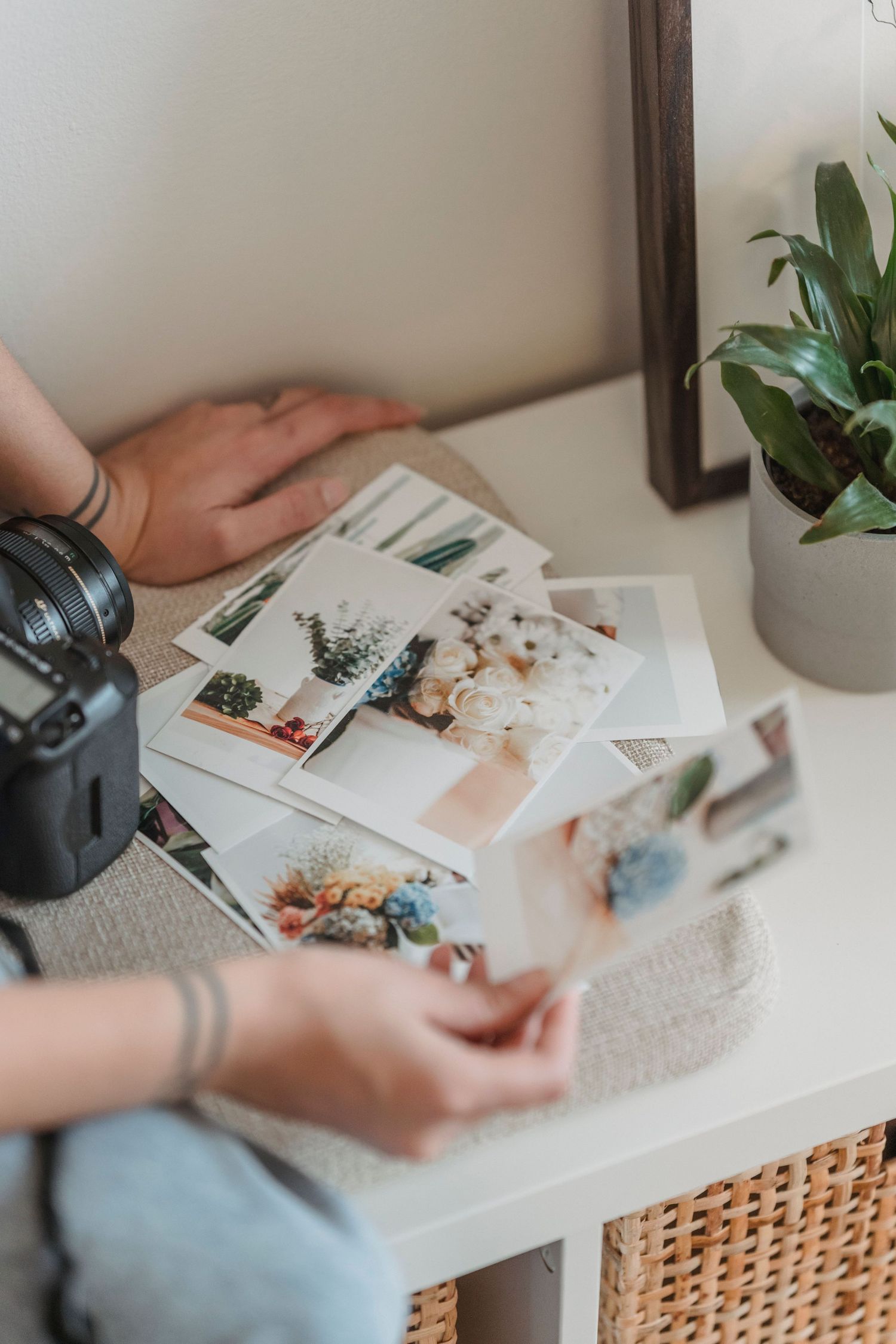 Person with tattoos reviewing printed photos on a white shelf beside a camera and green plant.