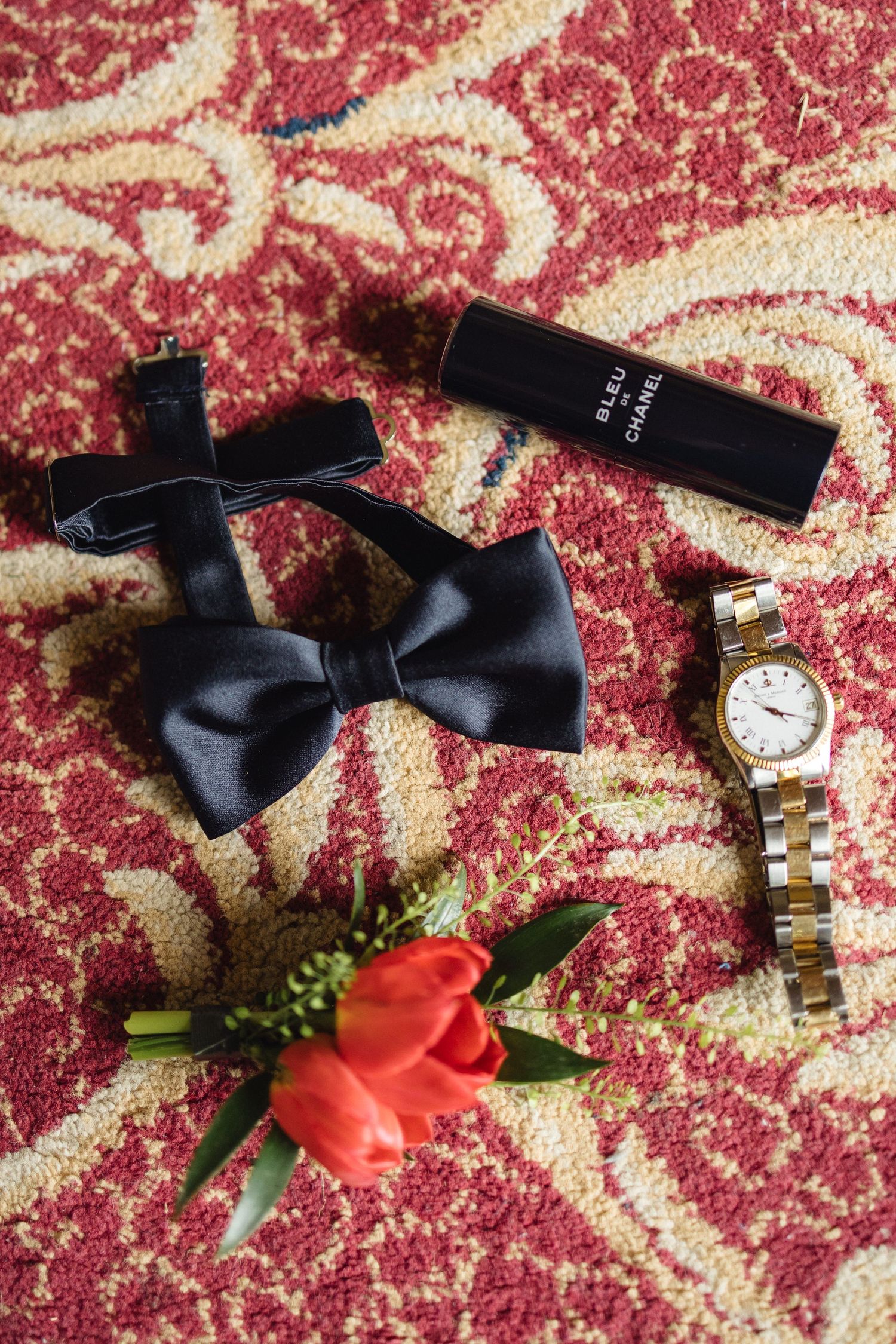 Groom wedding flat lay with black bow tie, Bleu de Chanel cologne, gold watch, and red tulip boutonniere.